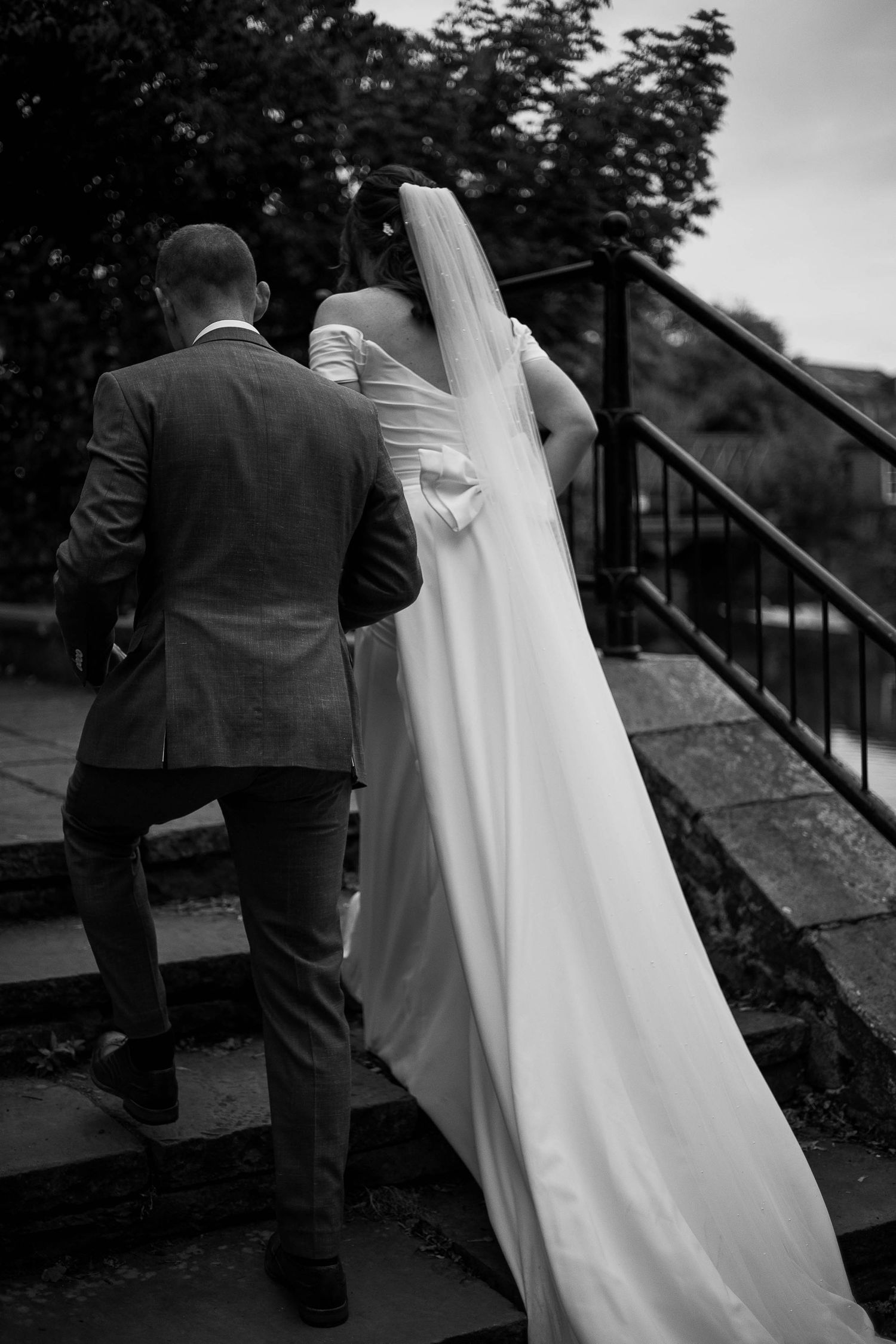 Black and white photo of bride and groom walking up stone steps, bride's long veil trailing behind her.