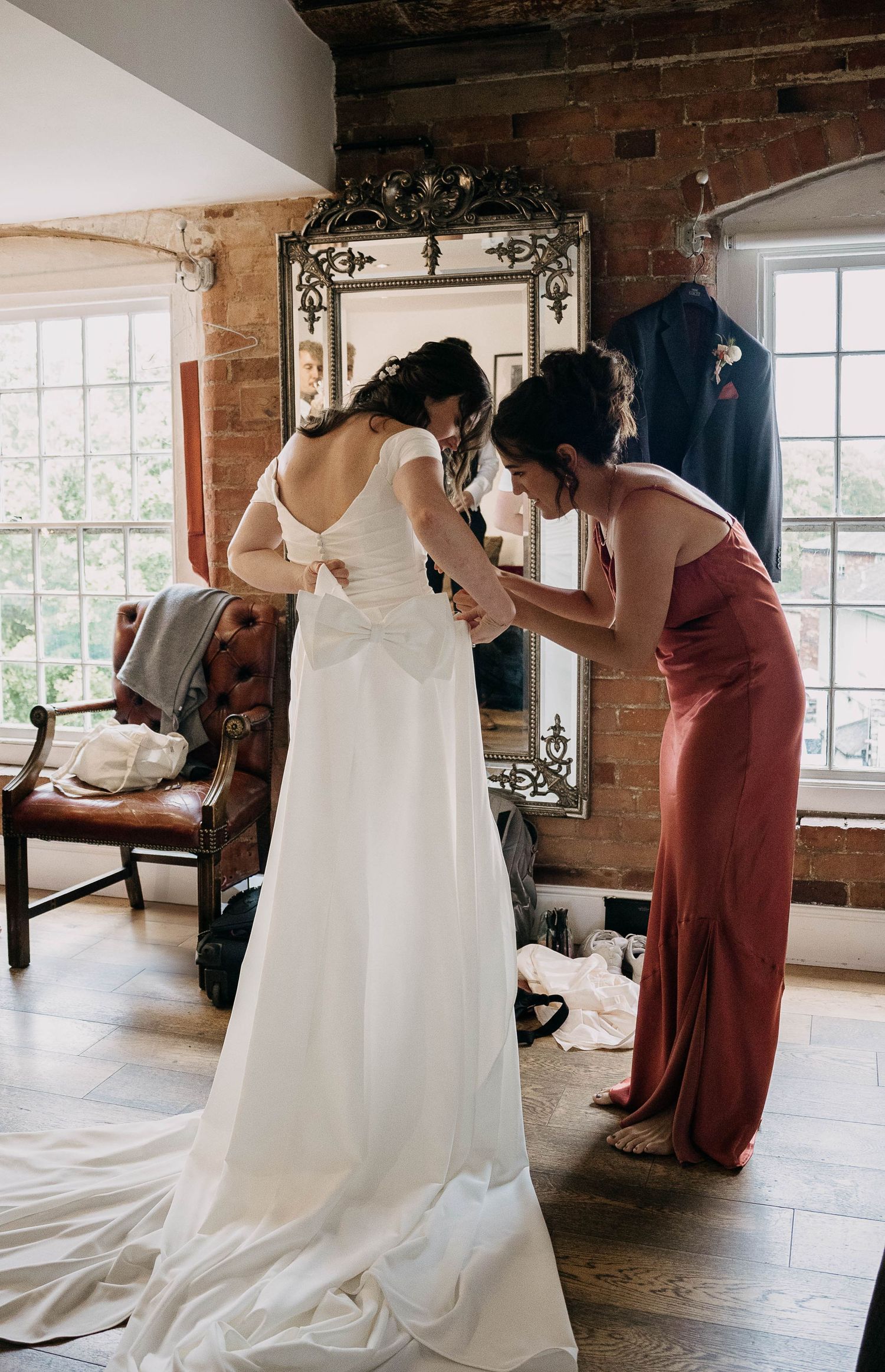 Bridesmaid in red dress fastening the back of a bride's white wedding gown in a rustic brick-walled room.