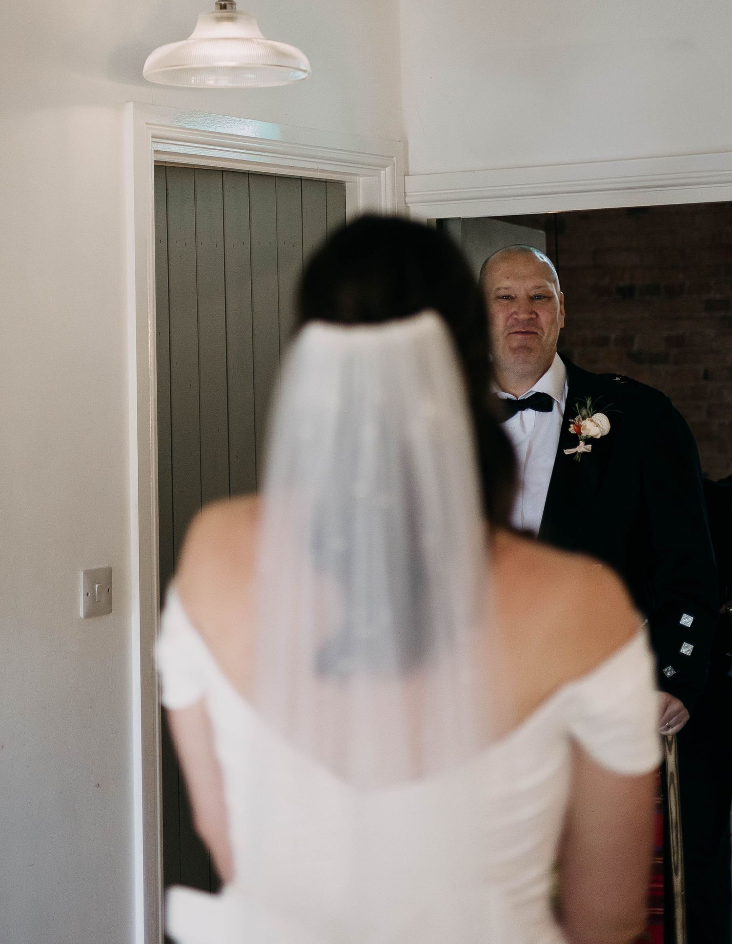 A bride in a white off-shoulder gown and veil shares a first look moment with a man in a black tuxedo.