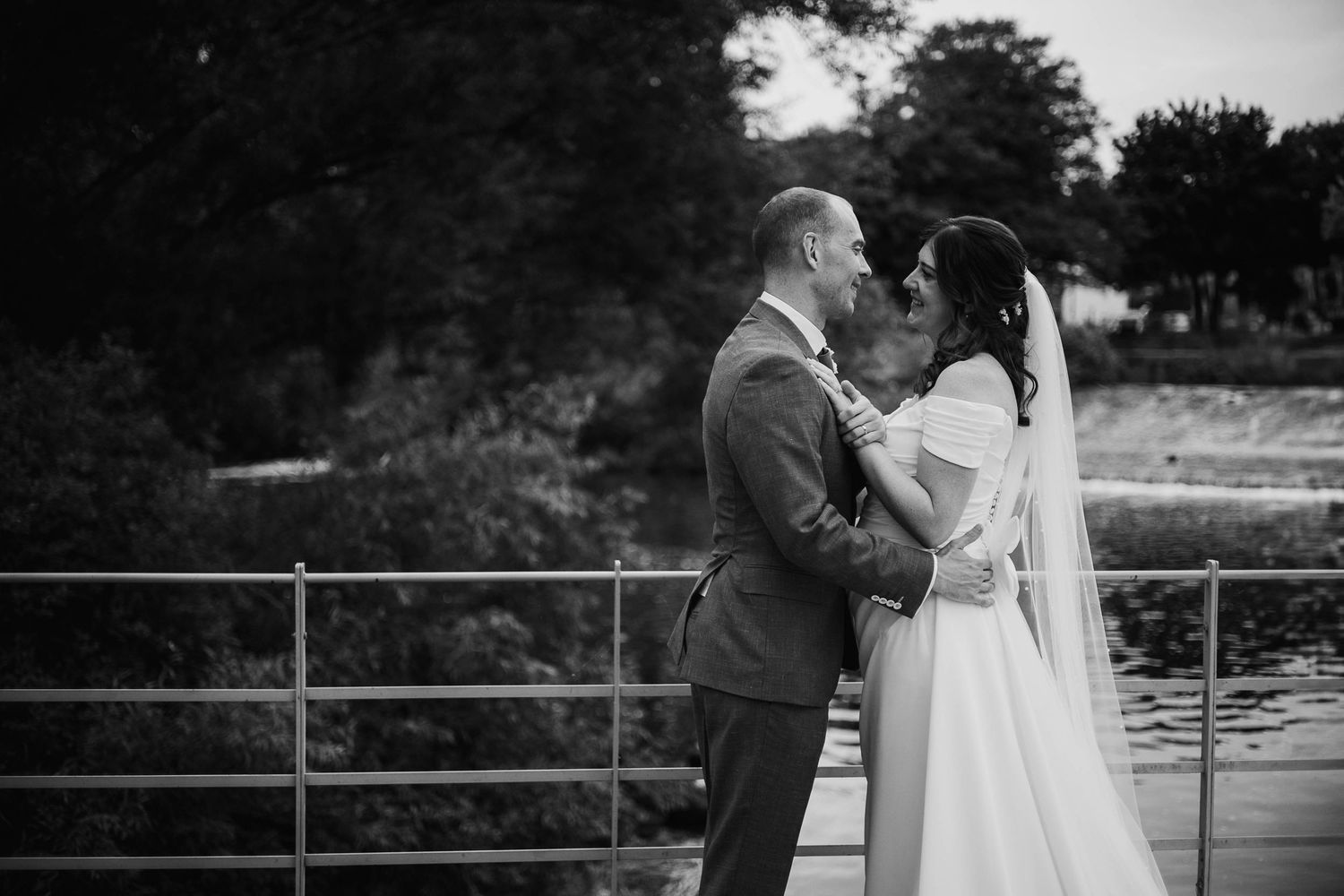 Black and white wedding photo of bride and groom embracing on a bridge near a serene river surrounded by lush trees.