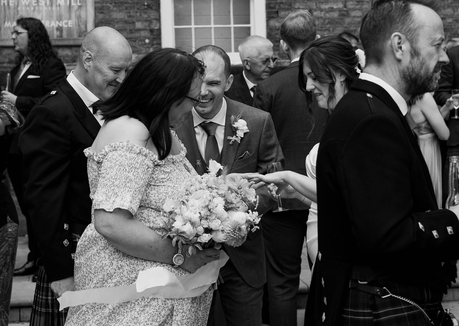 Black and white wedding photo of guests congratulating the bride holding a bouquet outside a stone building.