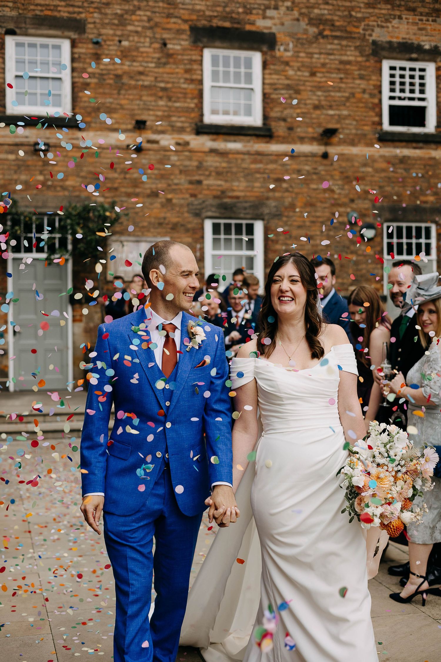Bride and groom walk through colorful confetti shower outside brick building, groom in blue suit, bride in white off-shoulder gown.