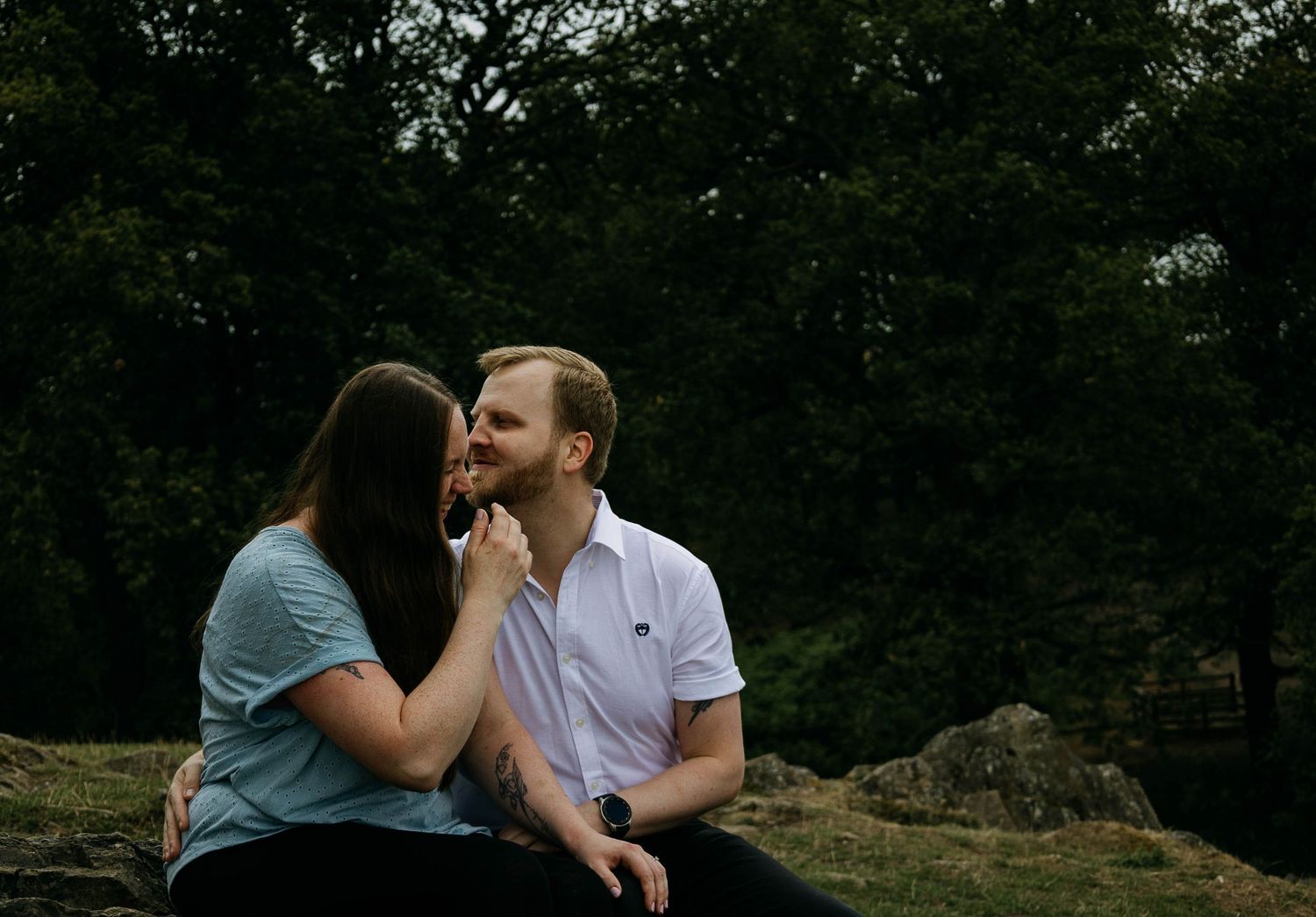Couple sitting outdoors on rocks, sharing a tender moment surrounded by lush green trees.