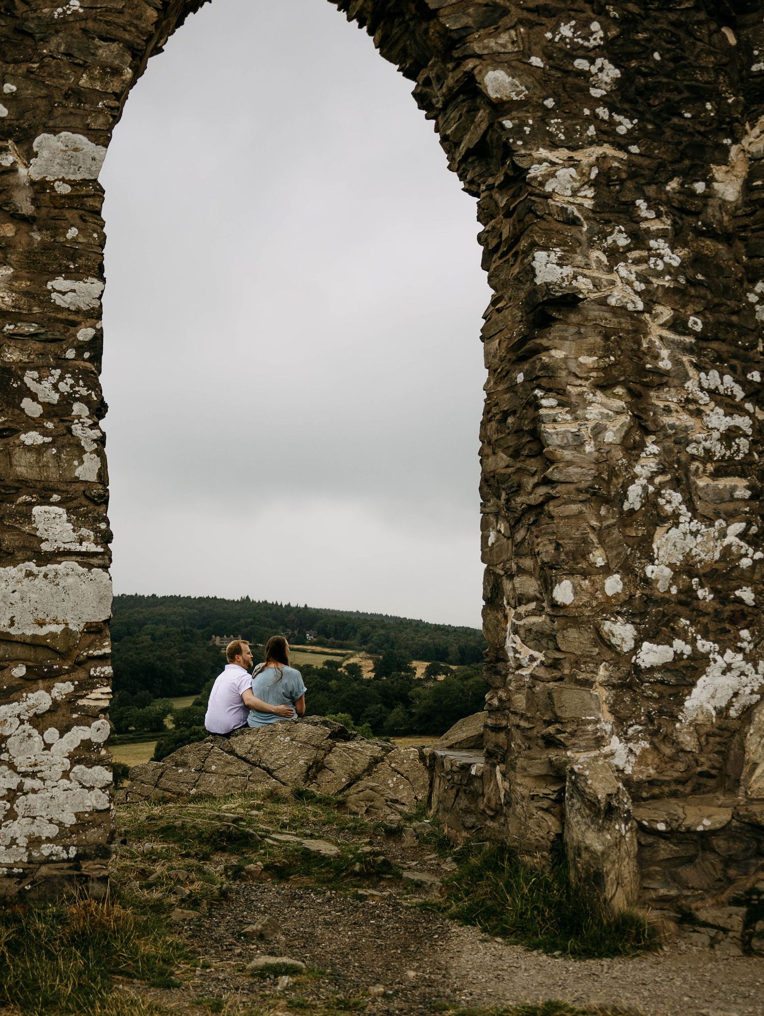 Couple sitting together framed by a ancient stone arch ruin overlooking a lush green valley on an overcast day.