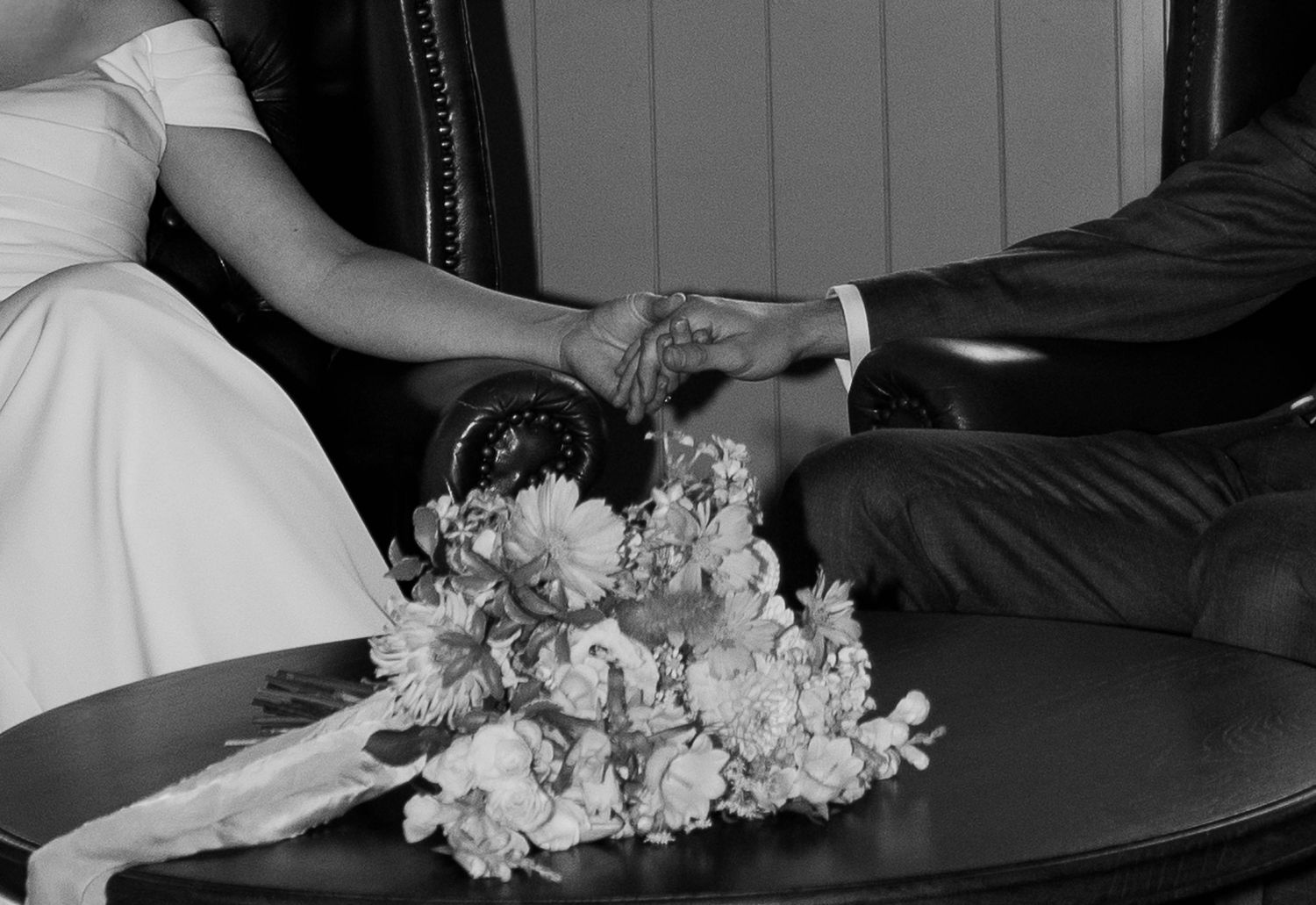 Black and white photo of bride and groom holding hands with a floral bouquet on a table.