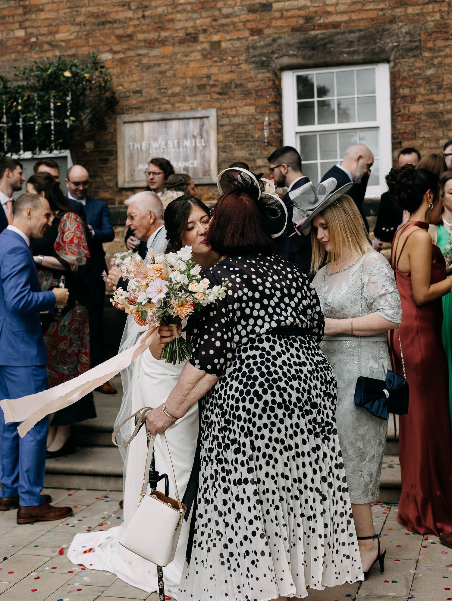 Bride in white gown hugging guests at outdoor wedding reception, colorful bouquet, brick building backdrop.