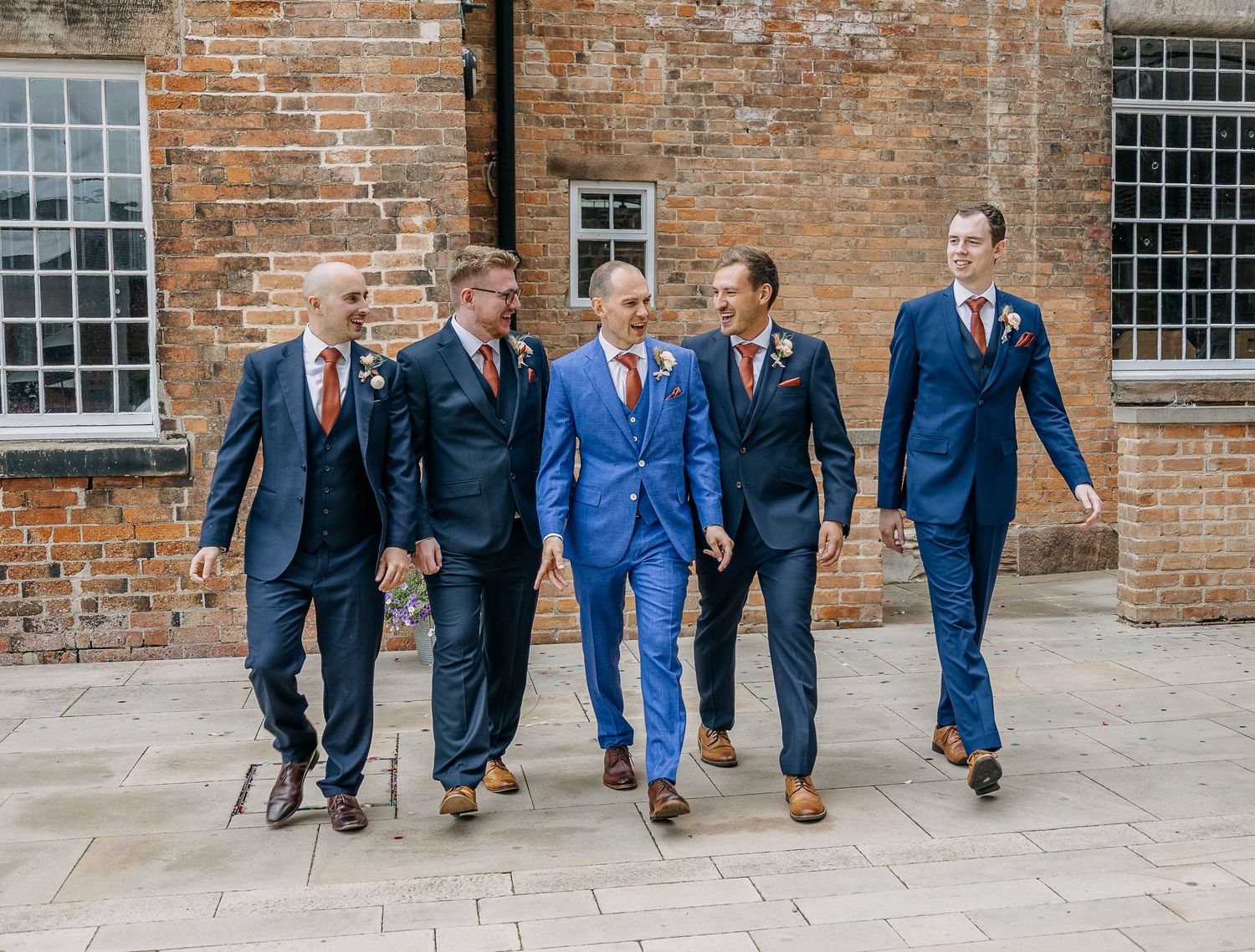 Groom in blue suit walks with four groomsmen in navy suits and red ties outside a brick building.