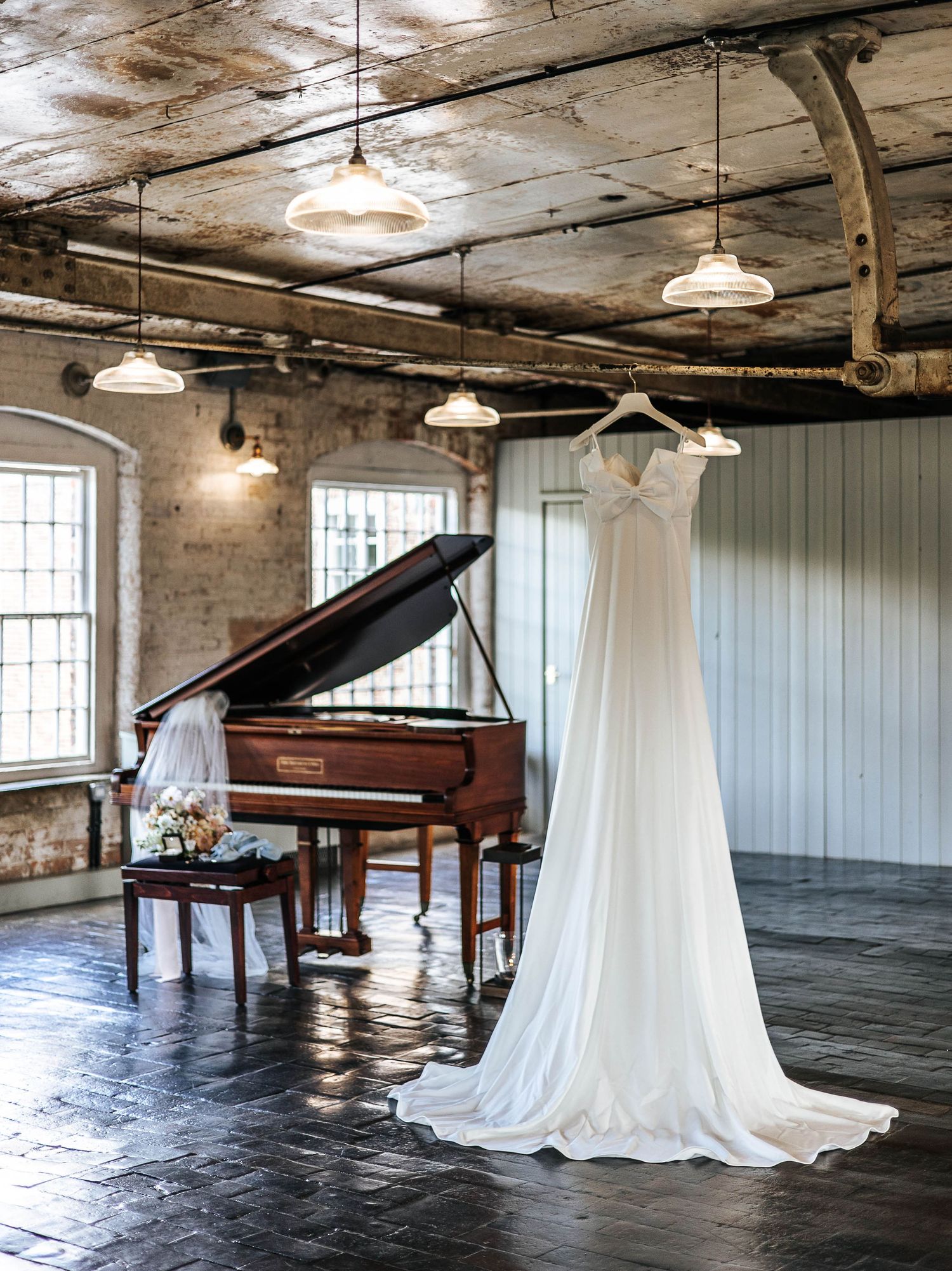 Elegant wedding dress hanging in a rustic industrial venue beside a grand piano with pendant lights overhead.