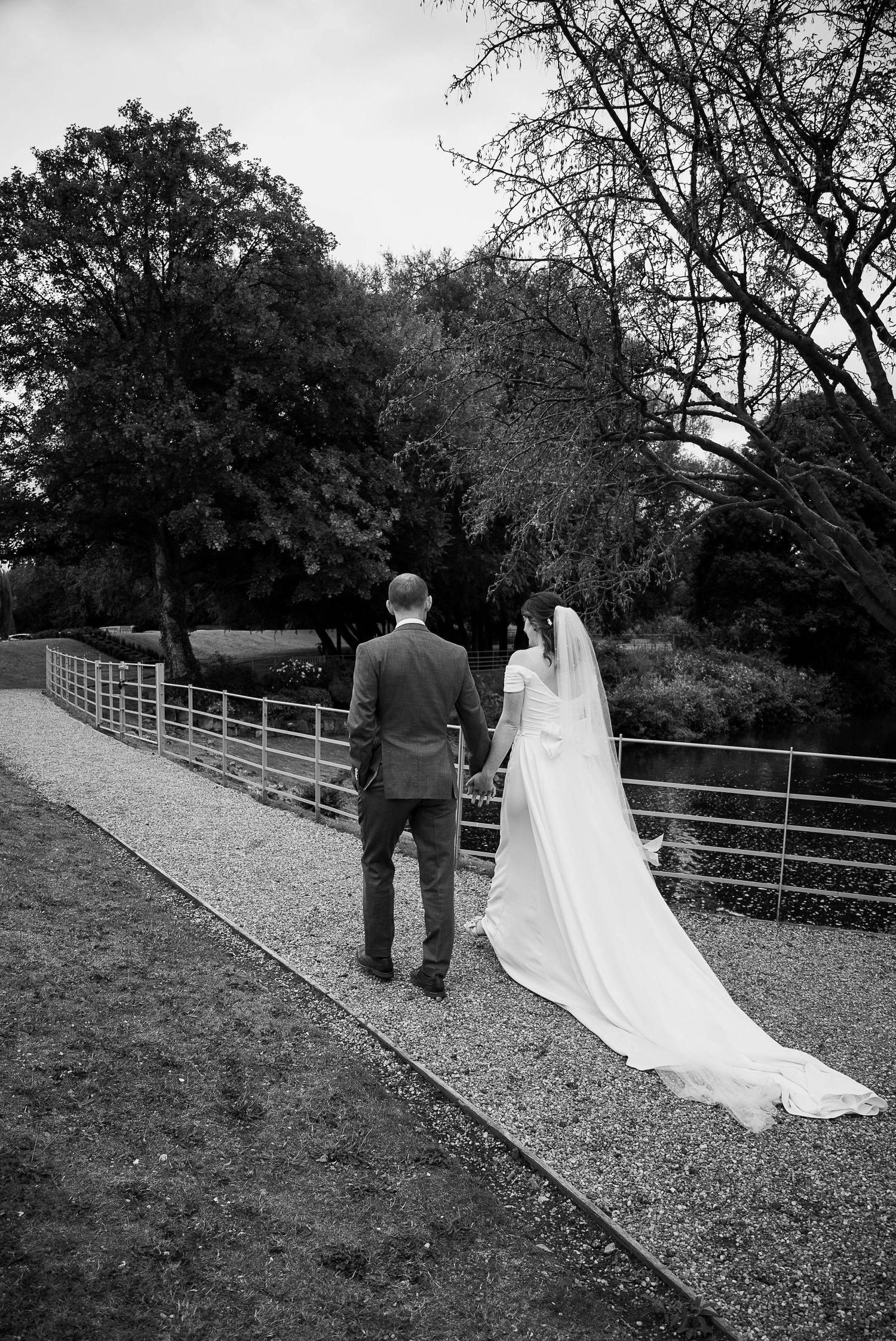 Black and white photo of a bride and groom walking hand in hand along a gravel path surrounded by trees.