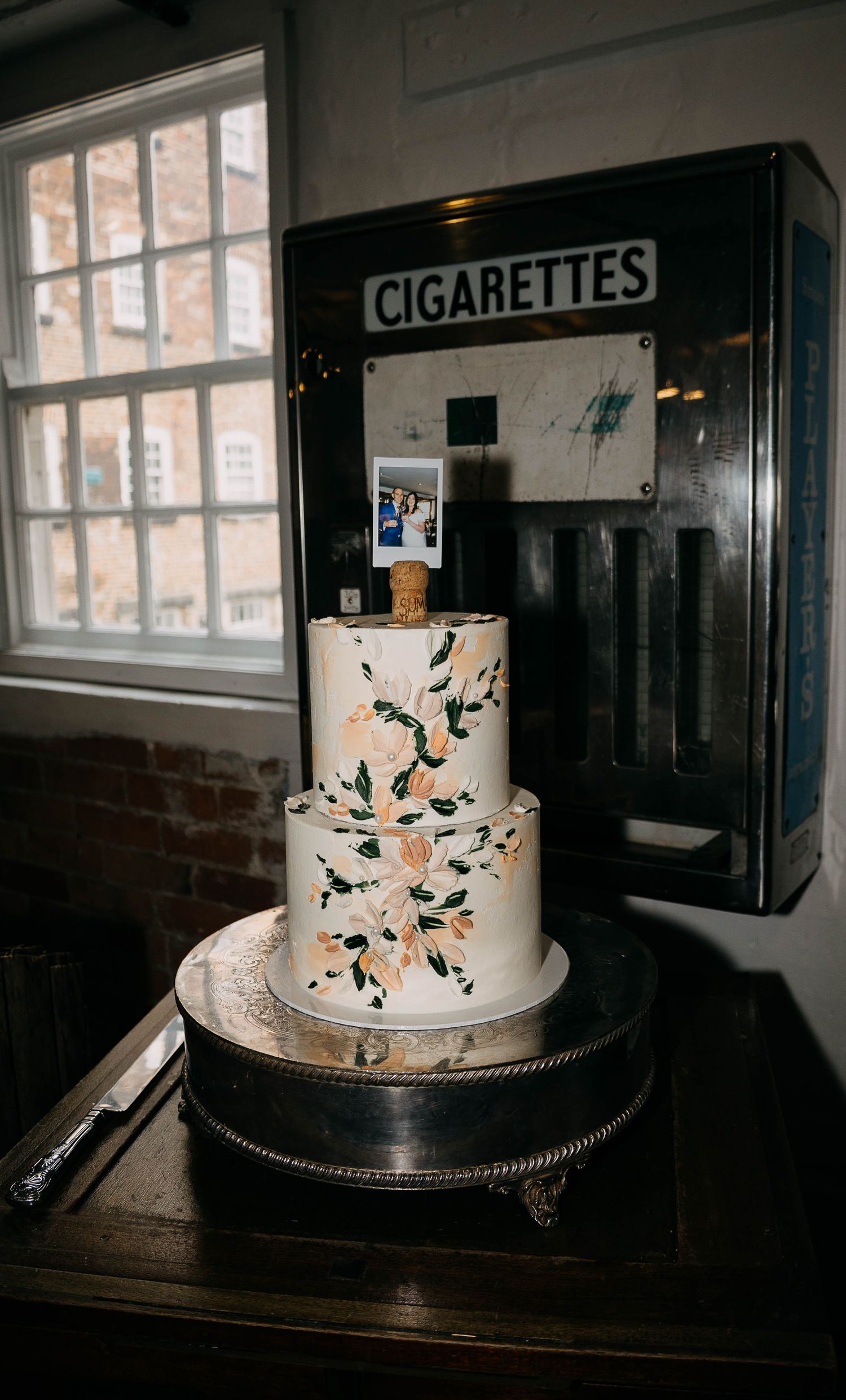 Two-tier floral wedding cake on silver stand displayed in front of vintage cigarette vending machine.