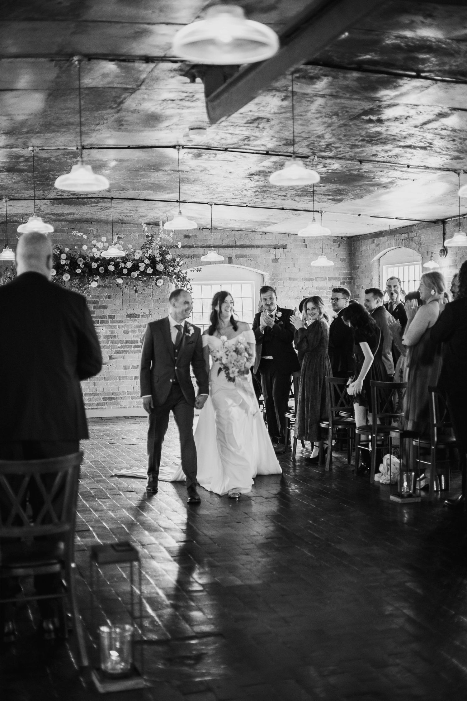 Black and white photo of a bride walking down the aisle with her escort in a rustic brick venue filled with wedding guests.