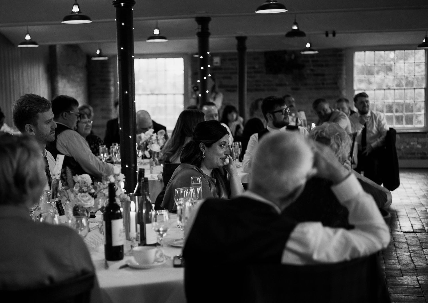 Black and white photo of elegantly dressed wedding guests dining at a candlelit reception in a rustic venue.