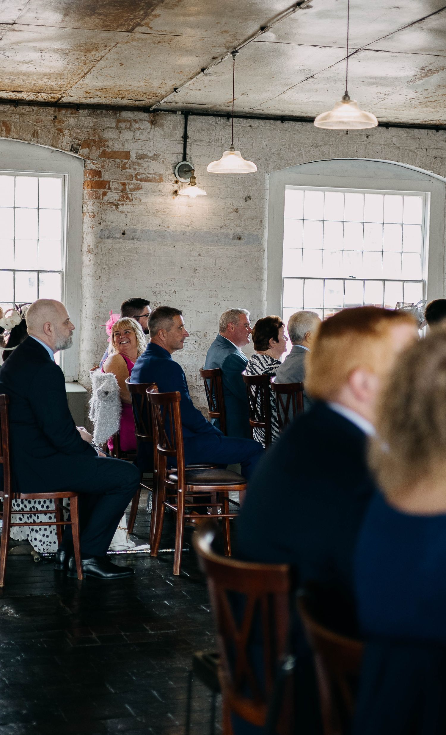 Wedding guests seated in rustic brick venue with pendant lights during an intimate ceremony.