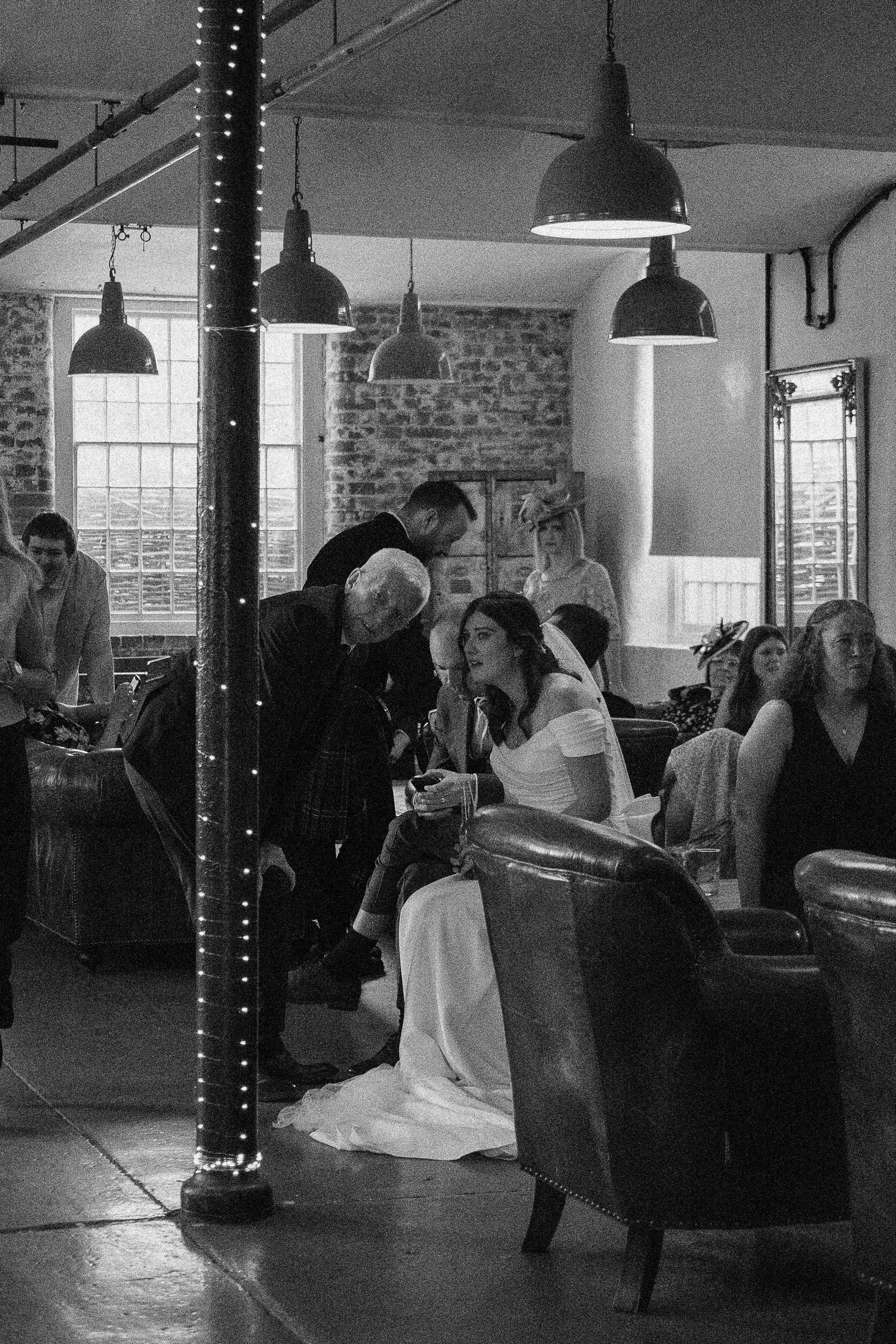 Black and white wedding photo of bride seated in armchair chatting with guests in rustic venue with fairy light pillar.