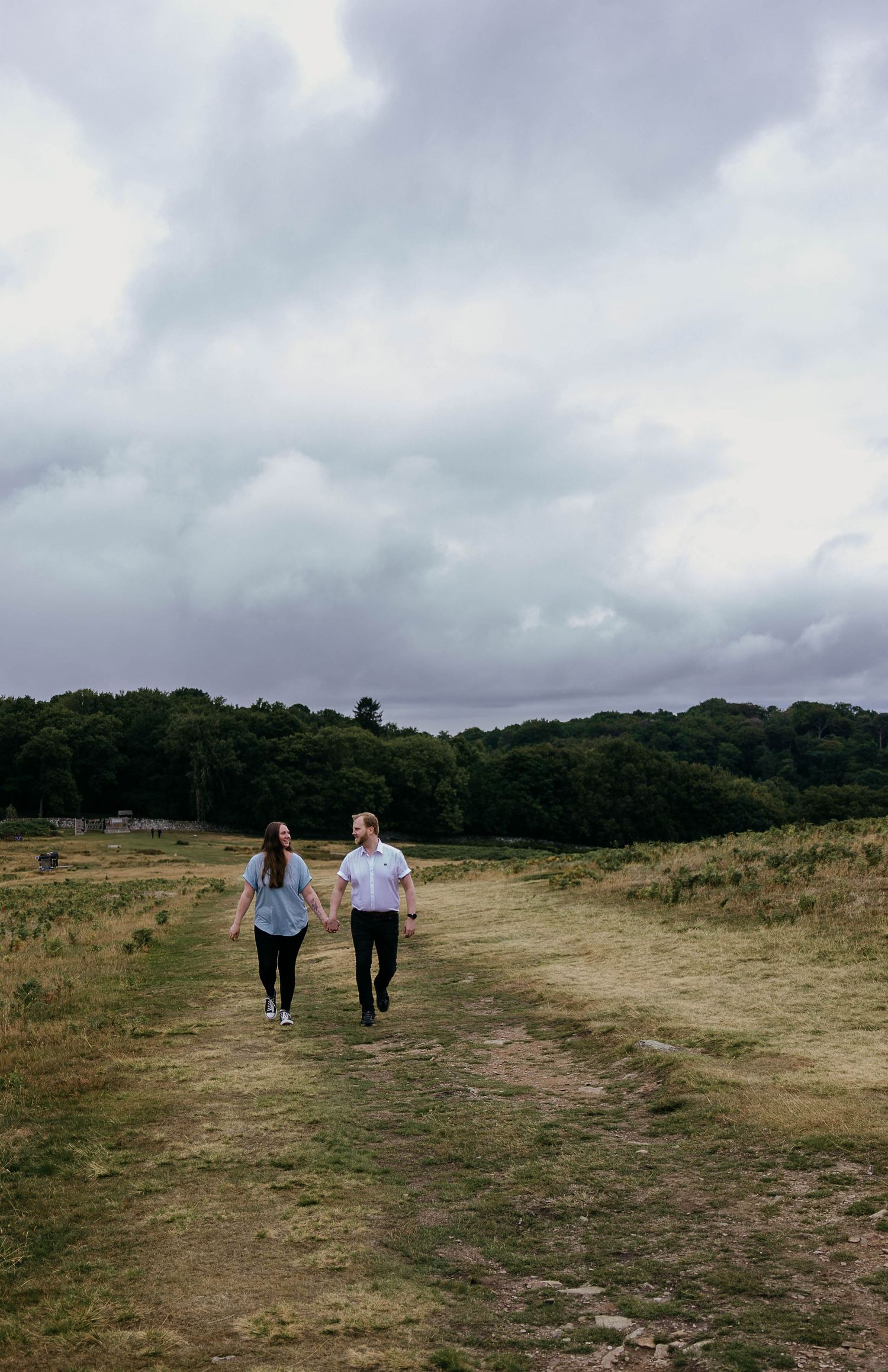 Couple holding hands walking along a grassy path in an open field with trees and cloudy sky.