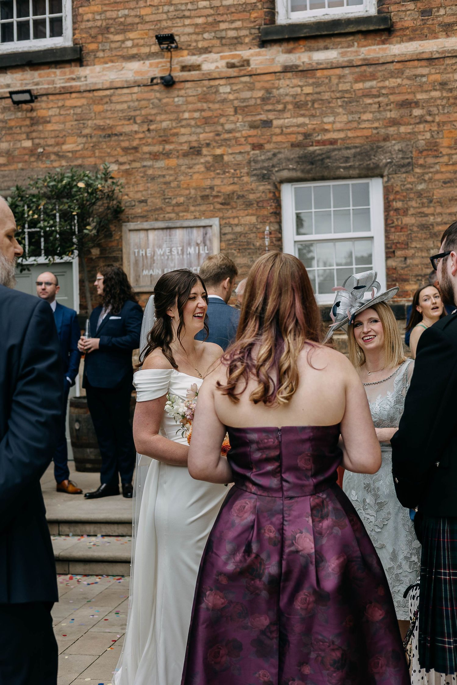 Bride in white off-shoulder gown chatting with guests in purple dress outside brick wedding venue.