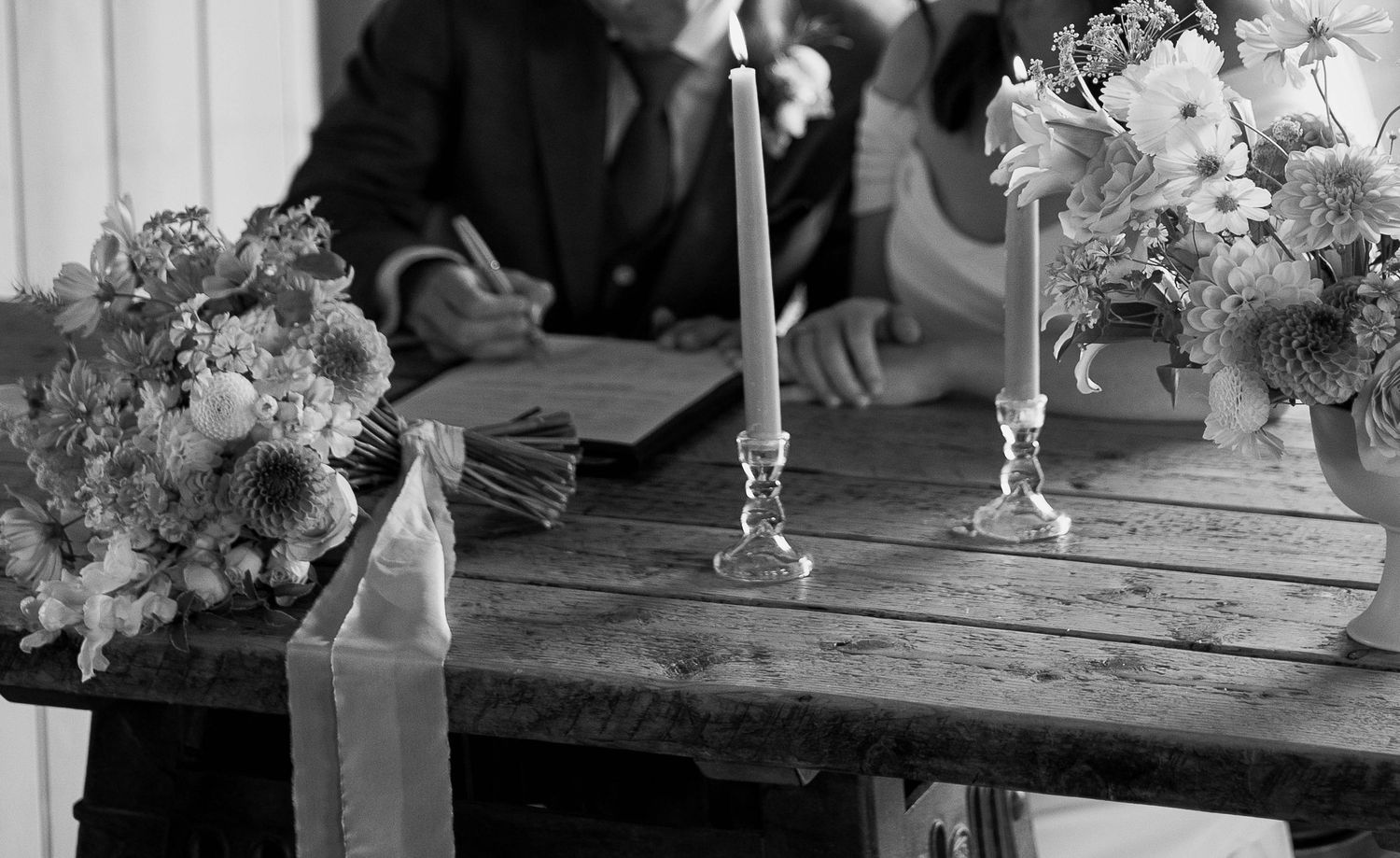 Black and white photo of a couple signing wedding documents at a rustic table with candles and floral arrangements.