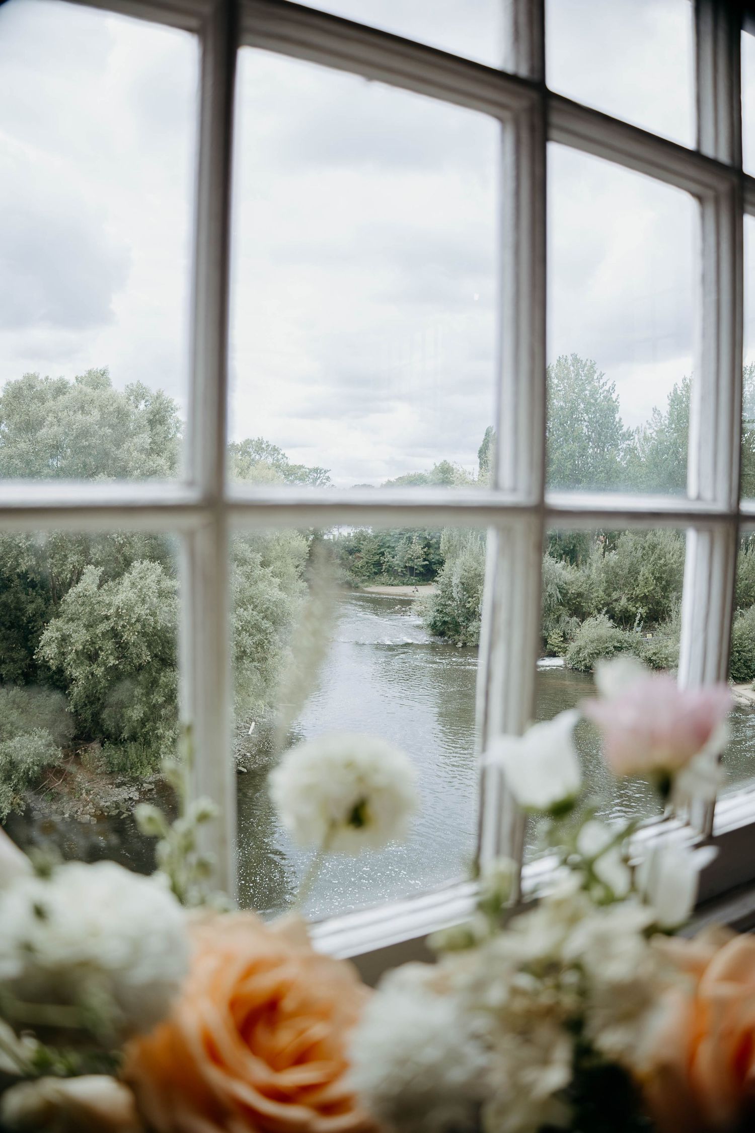 White-framed window overlooking a serene river and lush greenery, with soft peach and white roses in the foreground.