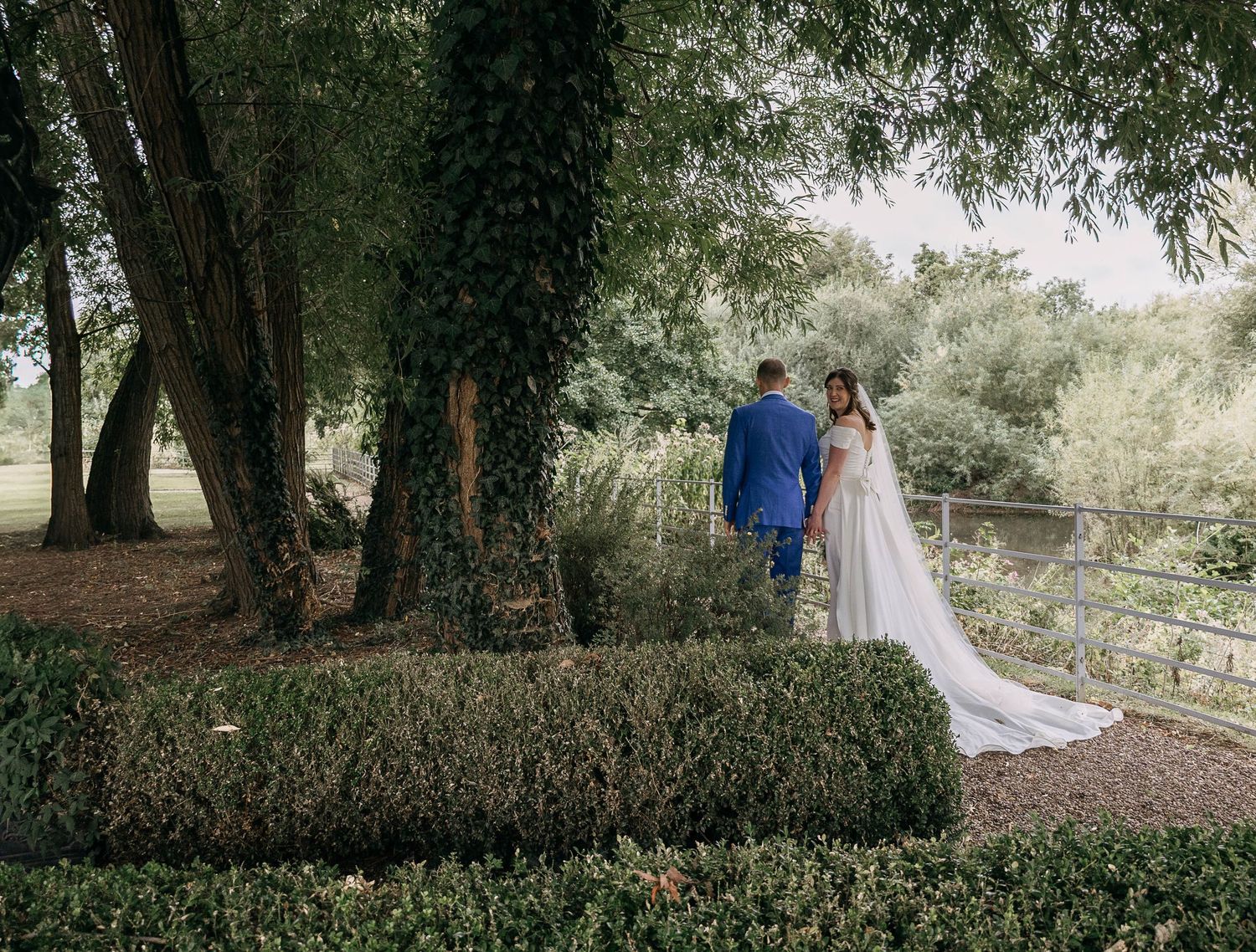 Bride in white gown and groom in blue suit stand among lush greenery and ivy-covered trees at outdoor wedding.