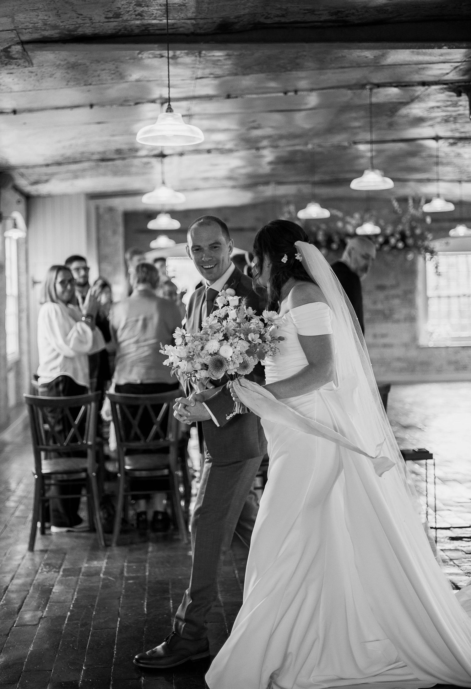 Black and white photo of a smiling groom walking with his bride in a wedding gown and veil inside a rustic venue.