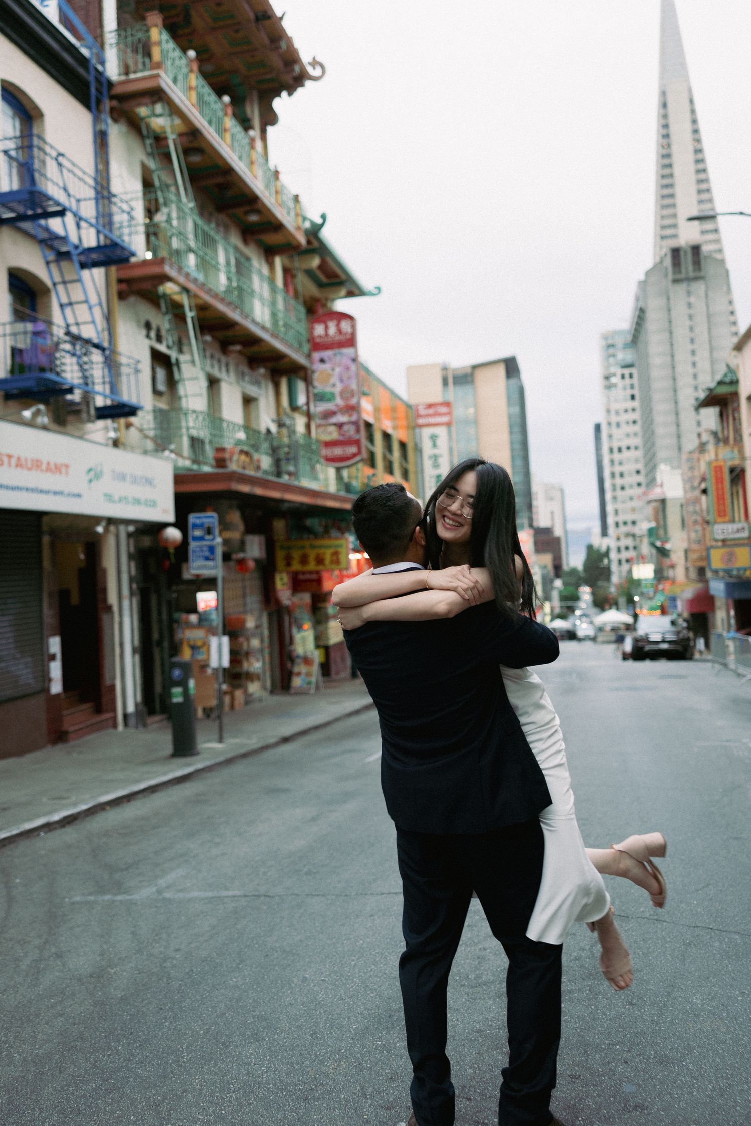 Two people dance playfully together on an empty city street with tall buildings and the Transamerica Pyramid in the background.