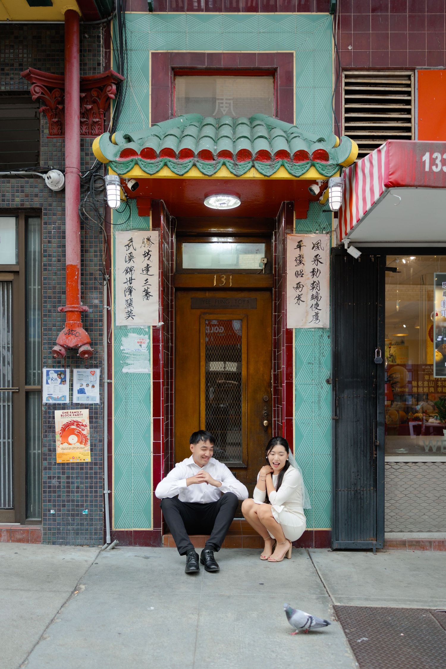 Two people sit in a doorway beneath a decorative green and turquoise awning in San Francisco's Chinatown while a pigeon walks by.