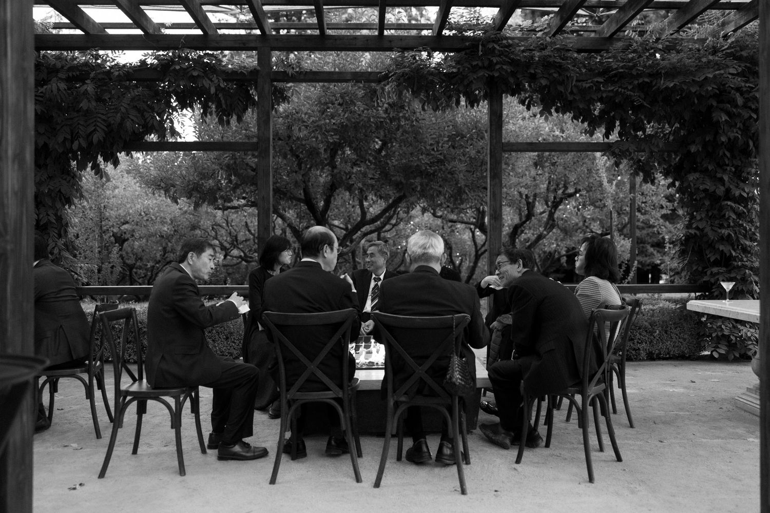 People sit at outdoor tables under a glass canopy in a garden setting with trees and natural light.