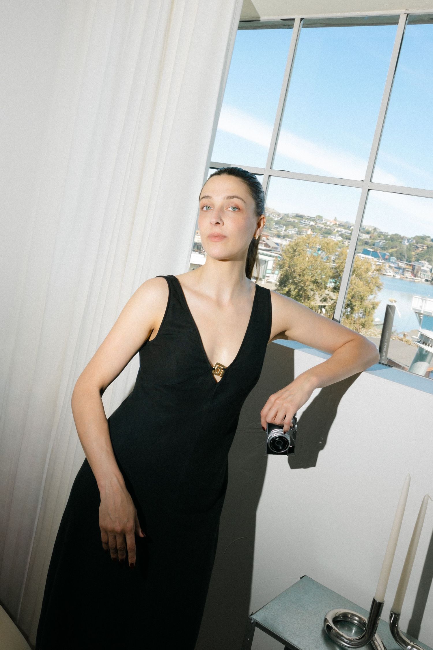 A person in a black sleeveless dress poses dramatically against a bright window with city views in the background.