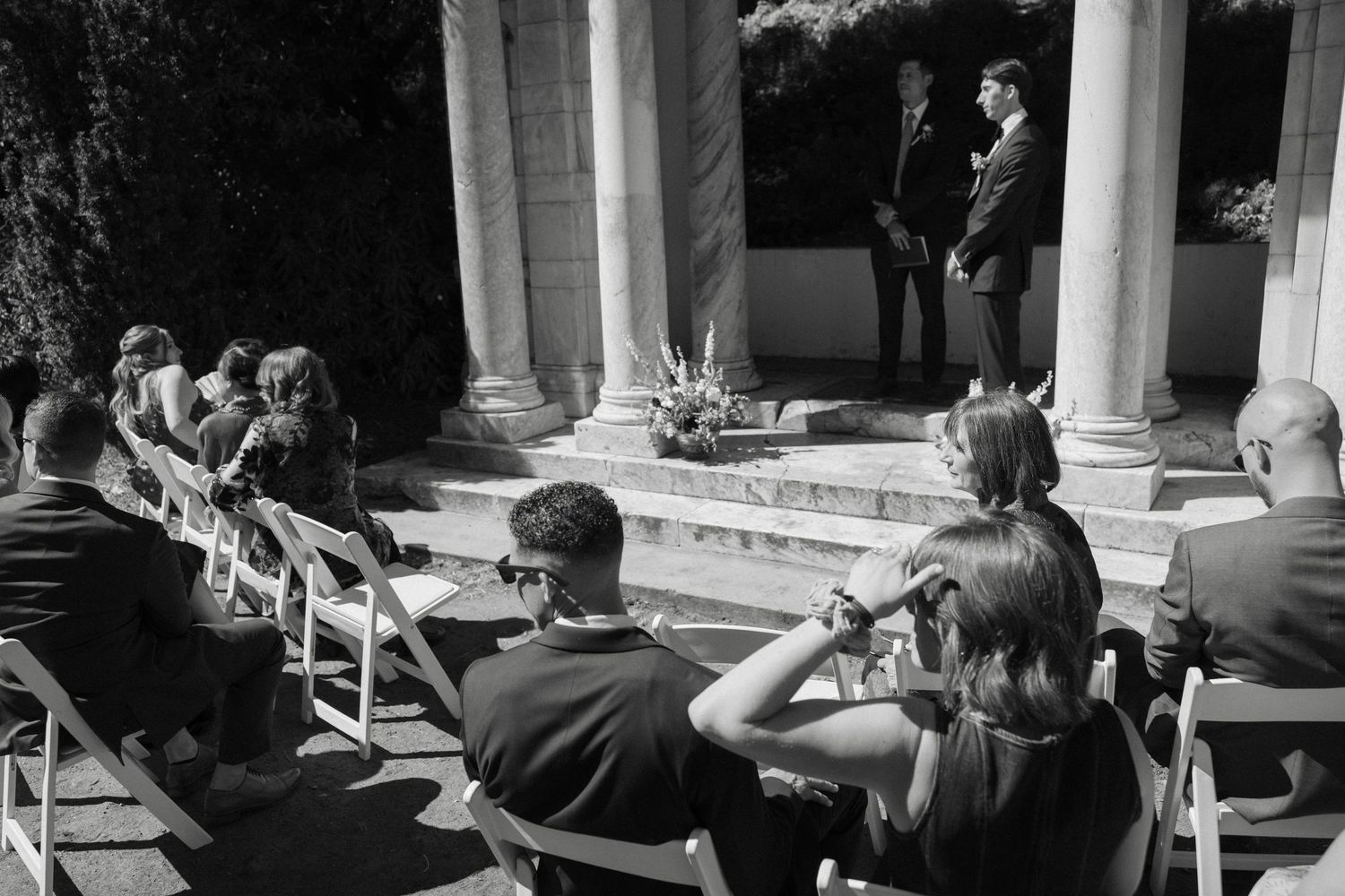 Black and white photo of an outdoor wedding ceremony with guests seated in chairs facing a columned gazebo structure.