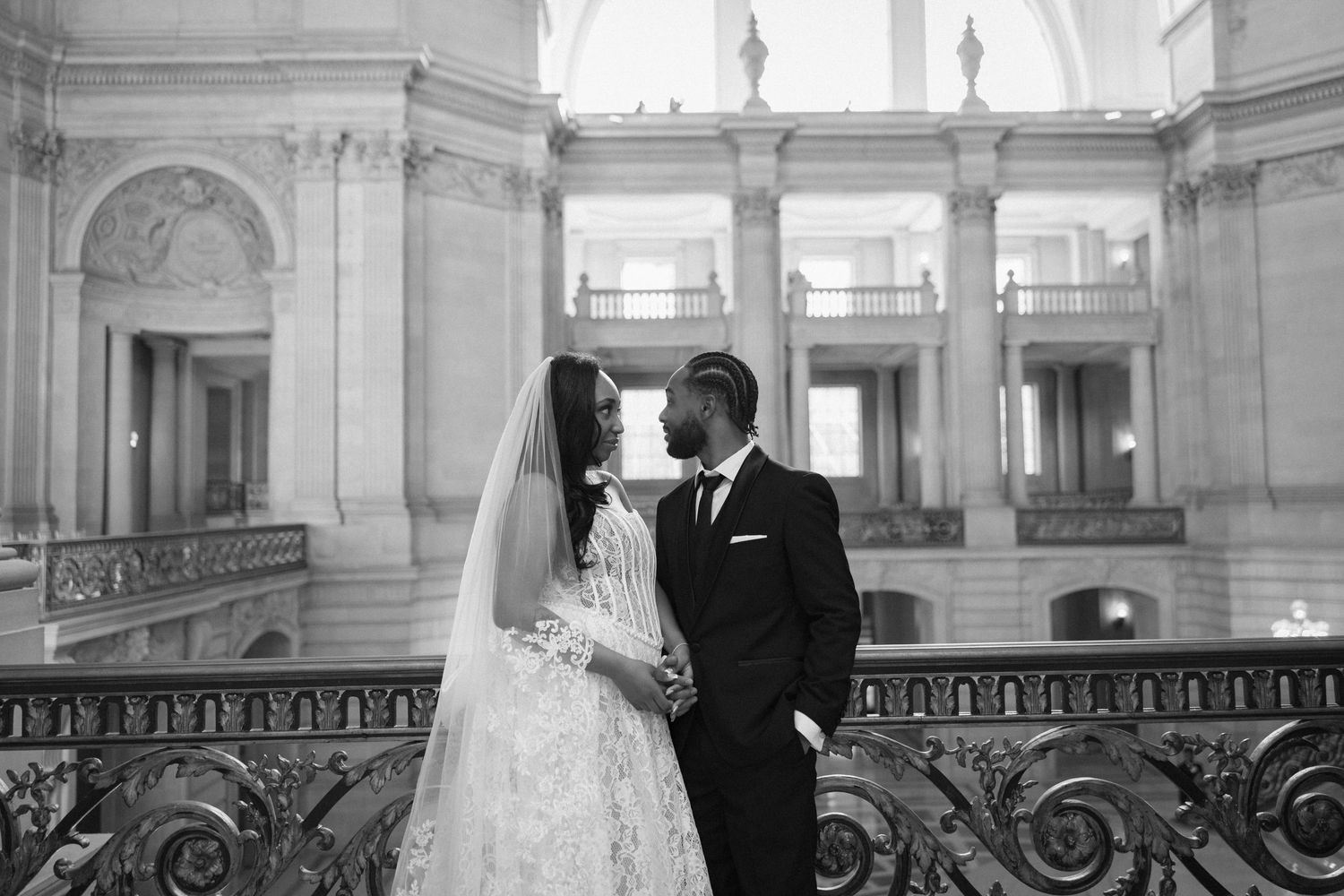 A bride and groom walk through an elegant white marble hallway with ornate architectural details and natural light.