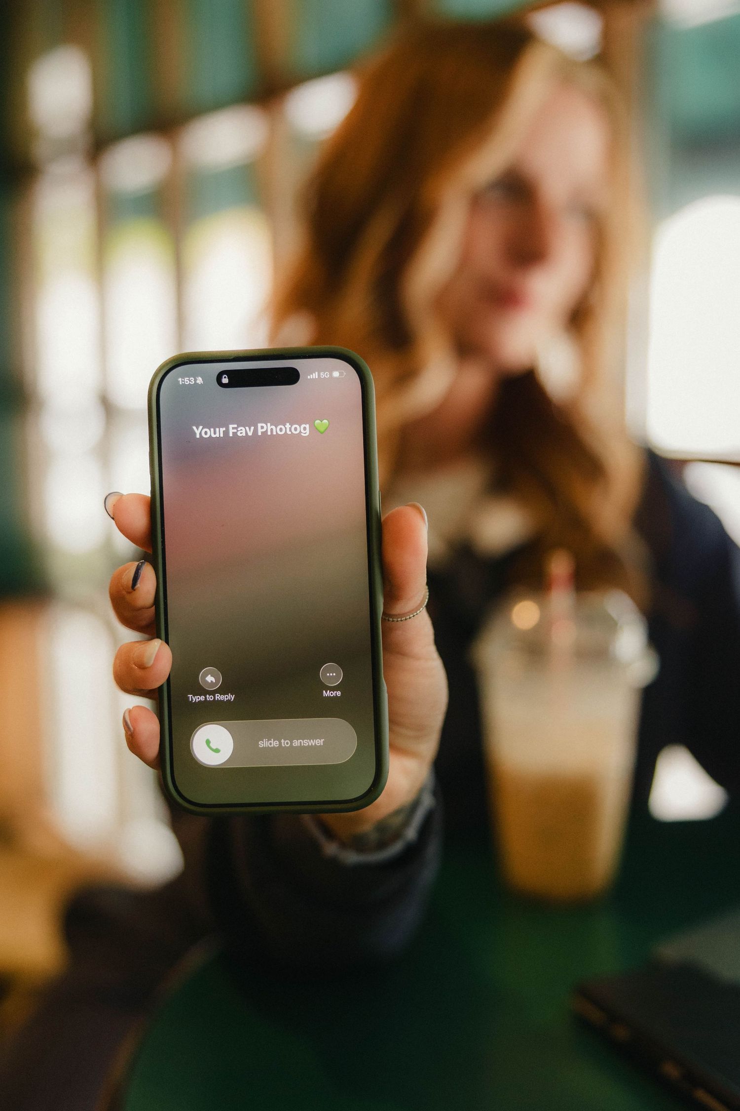 Woman in cafe holding iPhone showing incoming call screen labeled Your Fav Photog with iced coffee in background.