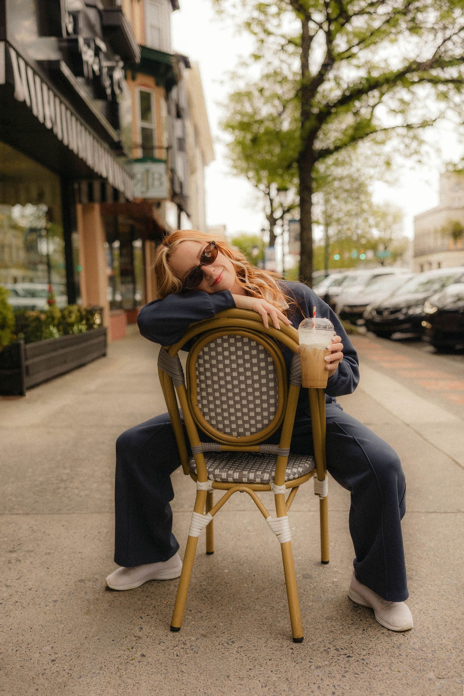 Woman in navy outfit sitting on bistro chair on urban sidewalk, holding iced coffee drink while wearing sunglasses.