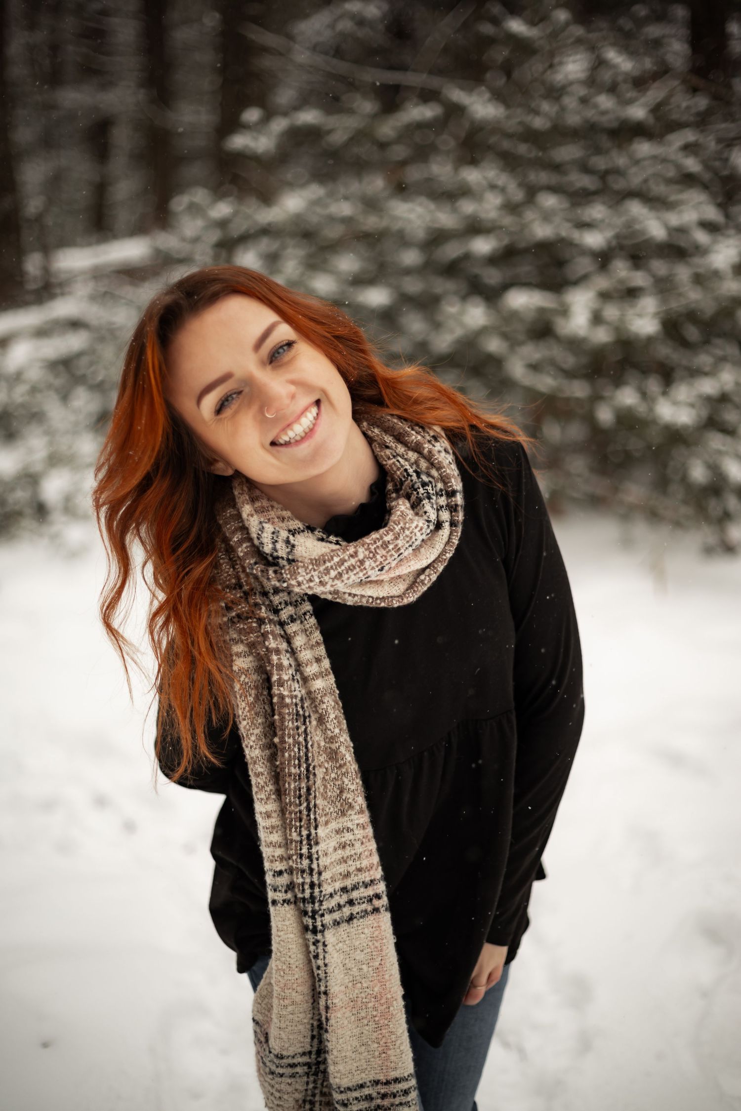 Woman with red hair wearing black coat and plaid scarf smiling in snowy winter setting.