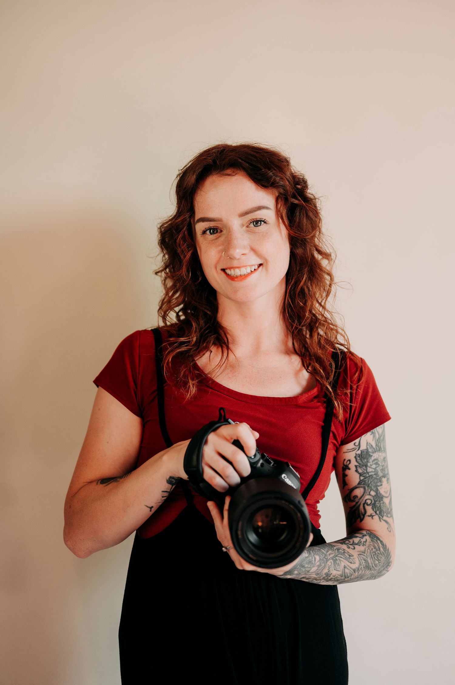 Smiling woman with curly hair holding camera, wearing red shirt against beige background.