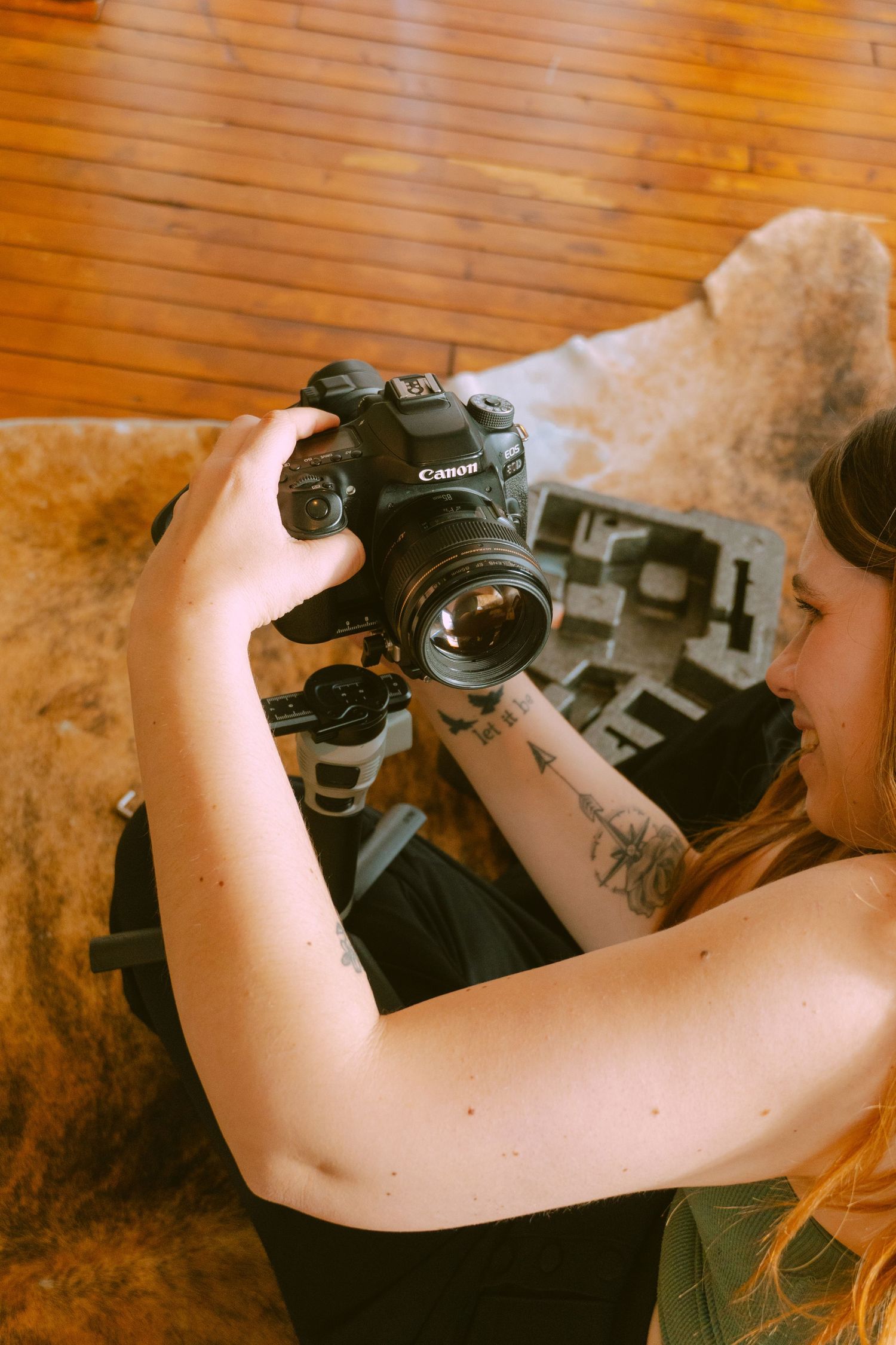 A person holds a Canon DSLR camera wearing black, showing a tattoo on their arm in a warm-toned indoor setting.