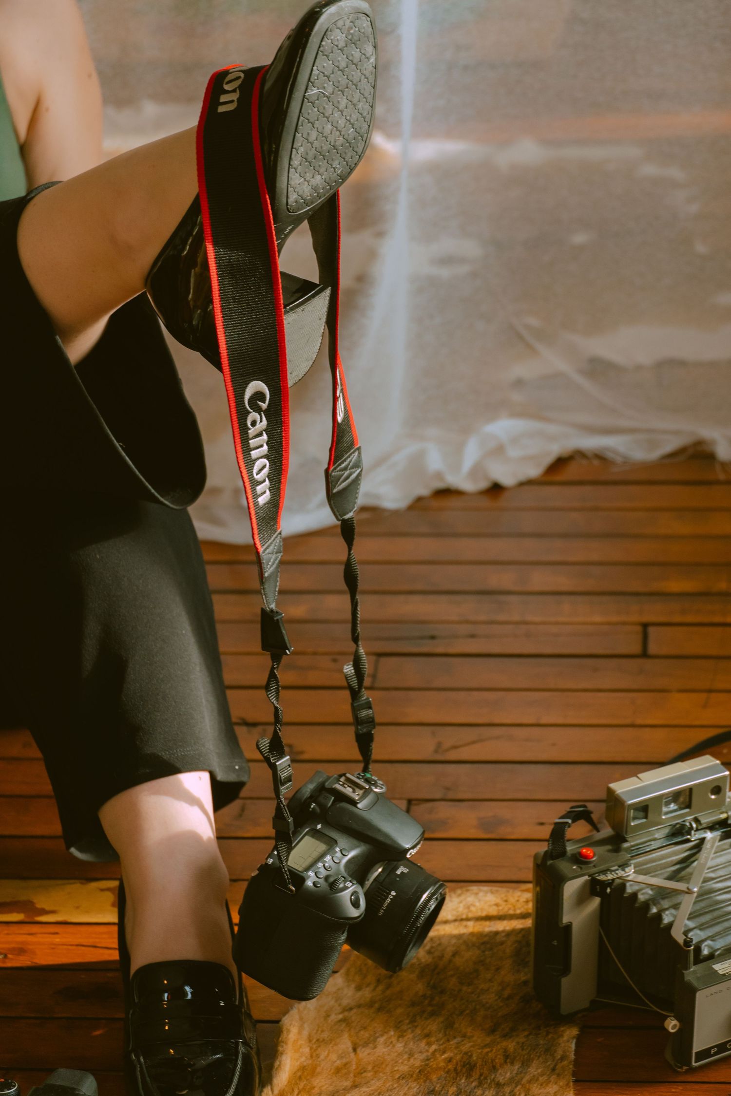 A Canon camera with red and black strap hangs near a pair of black shoes on a wooden floor.