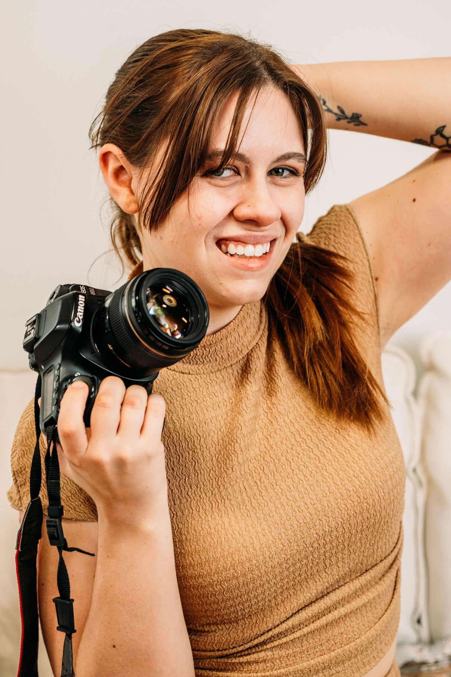 Photographer in tan sweater smiles while holding professional DSLR camera against light background.