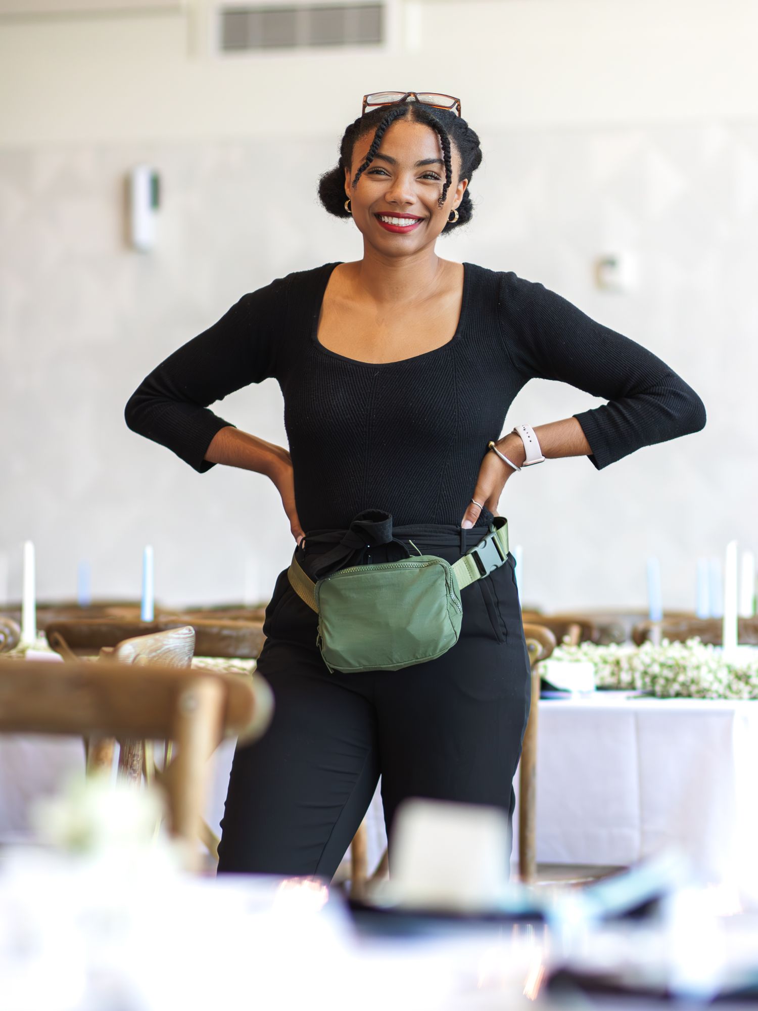 A hospitality worker in all black attire with a fanny pack smiles warmly while standing in a dining room setting.