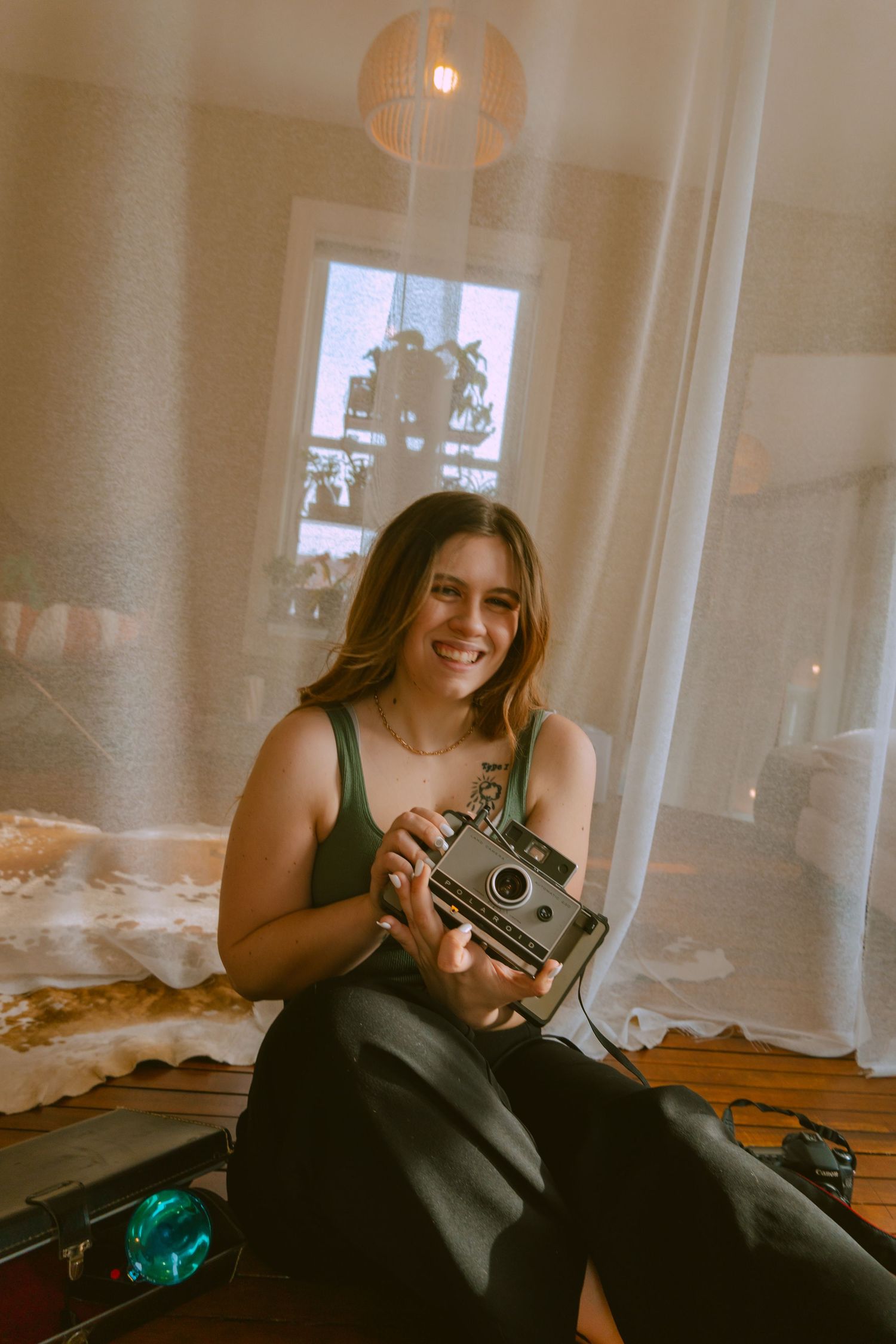 A person sits on a bed holding a vintage camera, with soft lighting and curtains in a cozy bedroom setting.