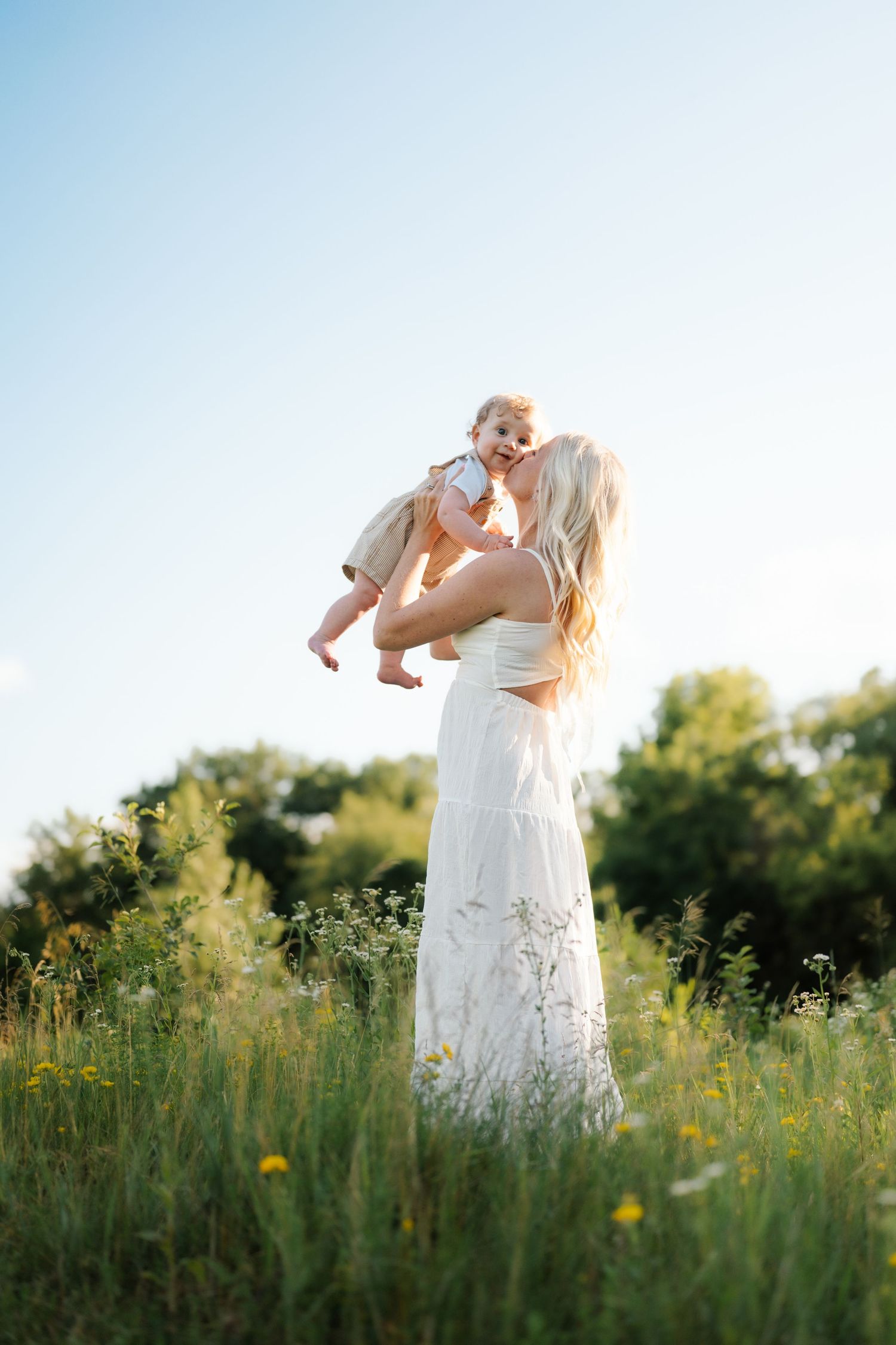 A mother in a white dress lifts her baby in a sunny wildflower field, sharing a joyful moment outdoors.