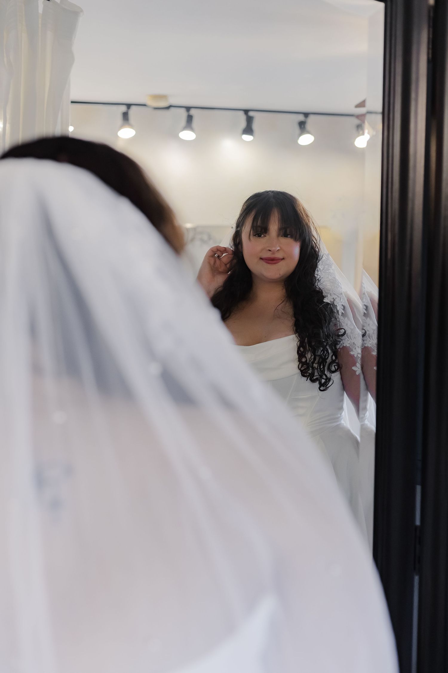 A bride in a white wedding dress stands in front of a mirror with blurred reflections creating an artistic effect.