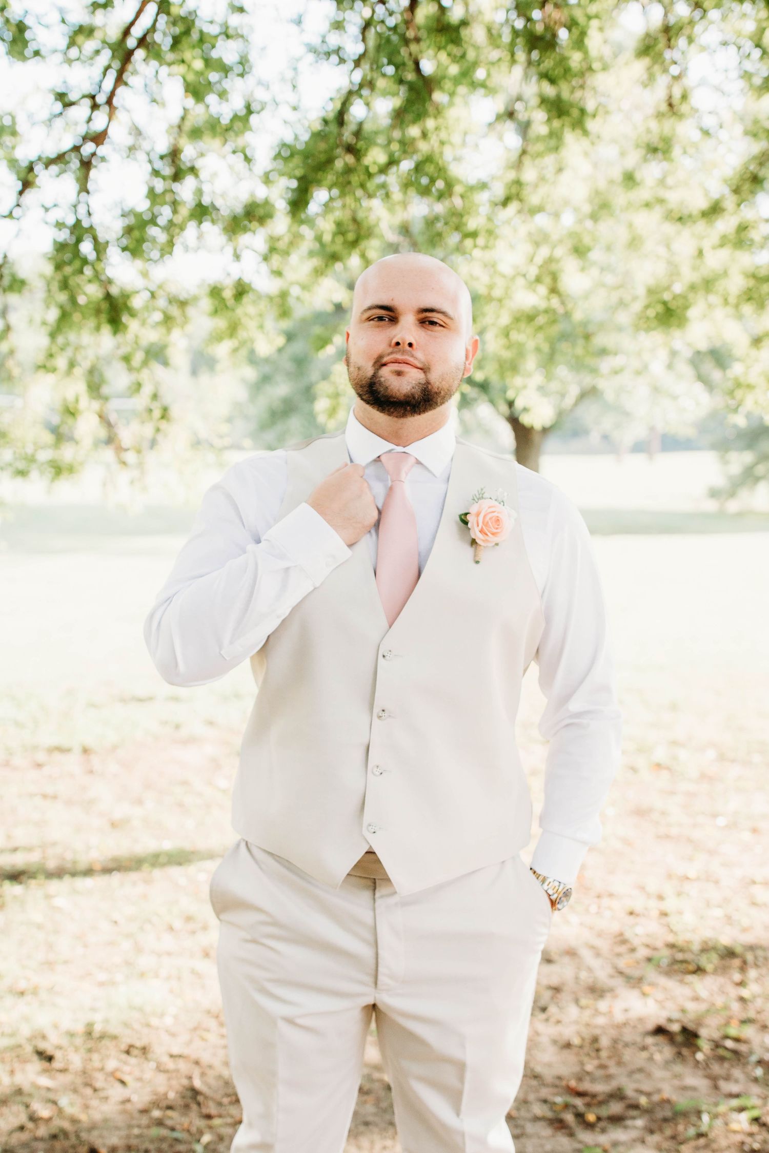 Groom in cream colored suit adjusting tie during outdoor wedding photoshoot.