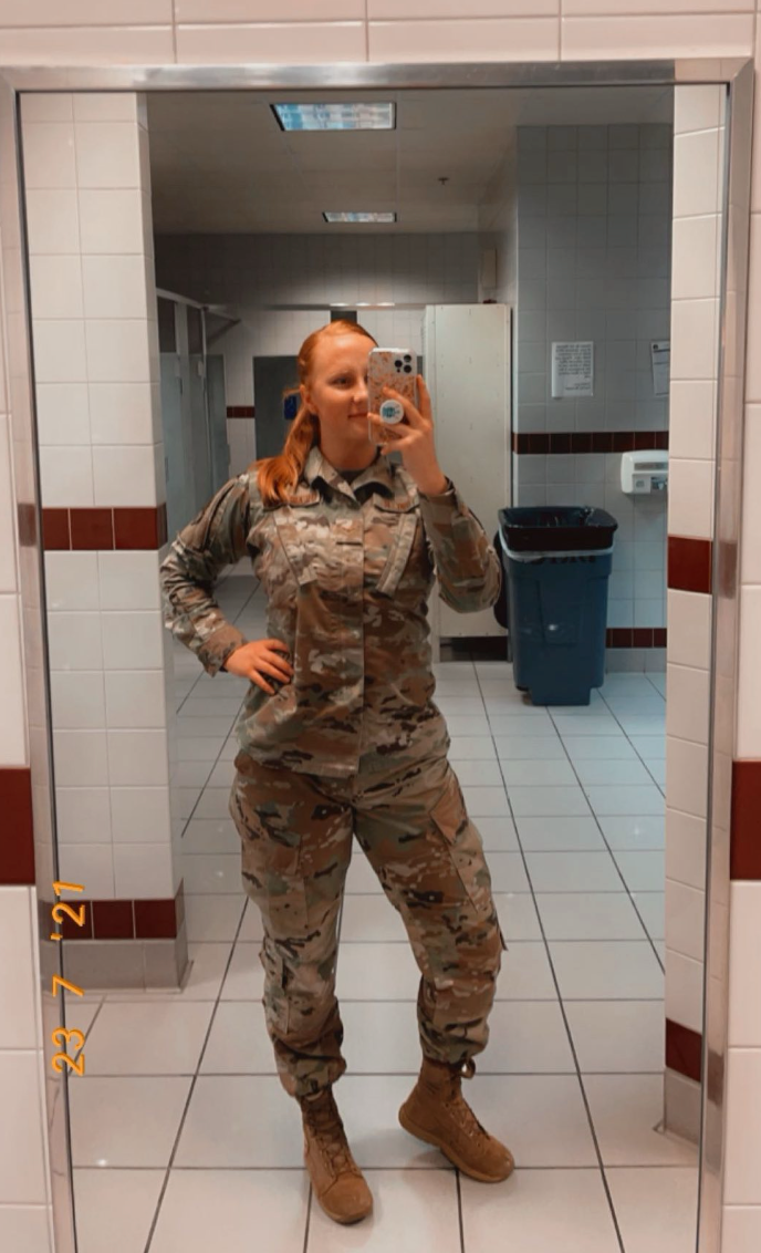 Military service member in camouflage uniform taking mirror selfie in hallway.