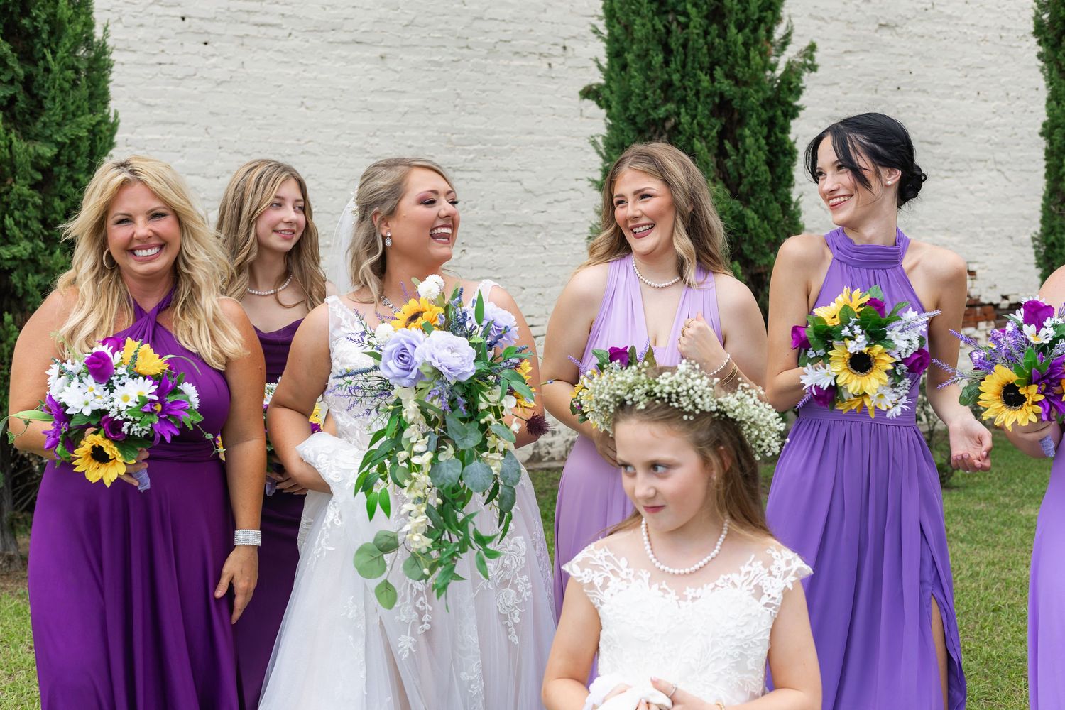Wedding party in purple bridesmaid dresses poses outdoors against white brick wall.
