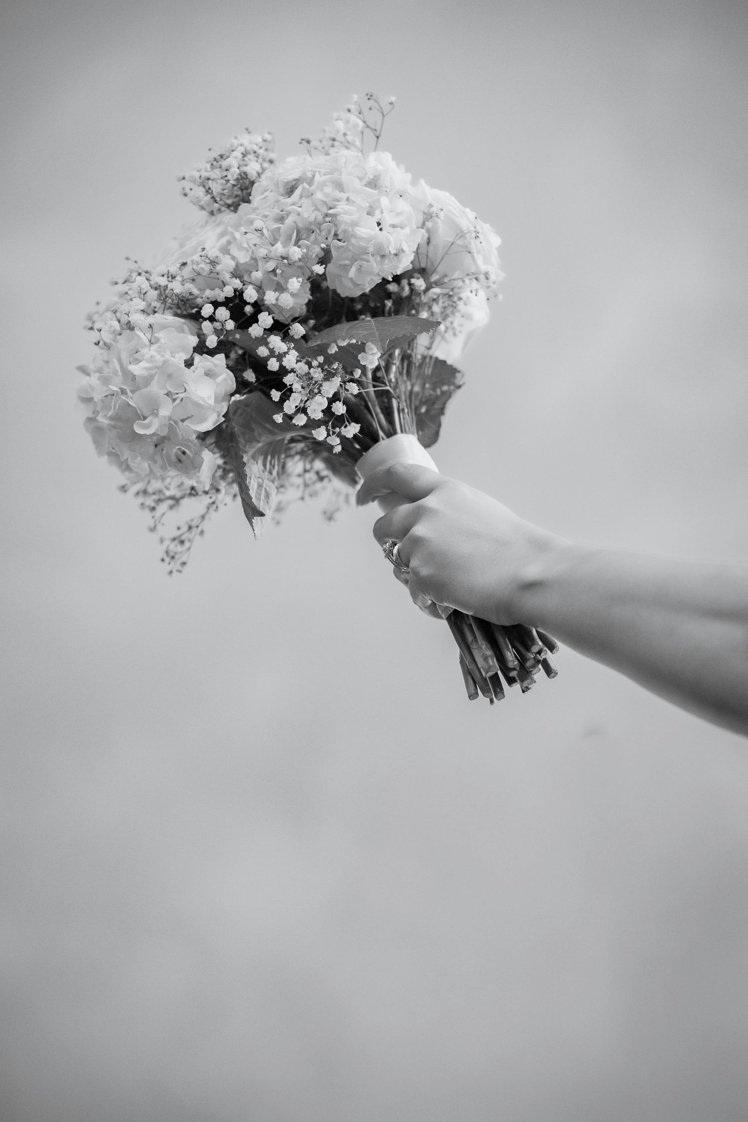 A hand holds a delicate bouquet of white flowers and baby's breath against a soft gray background in black and white.