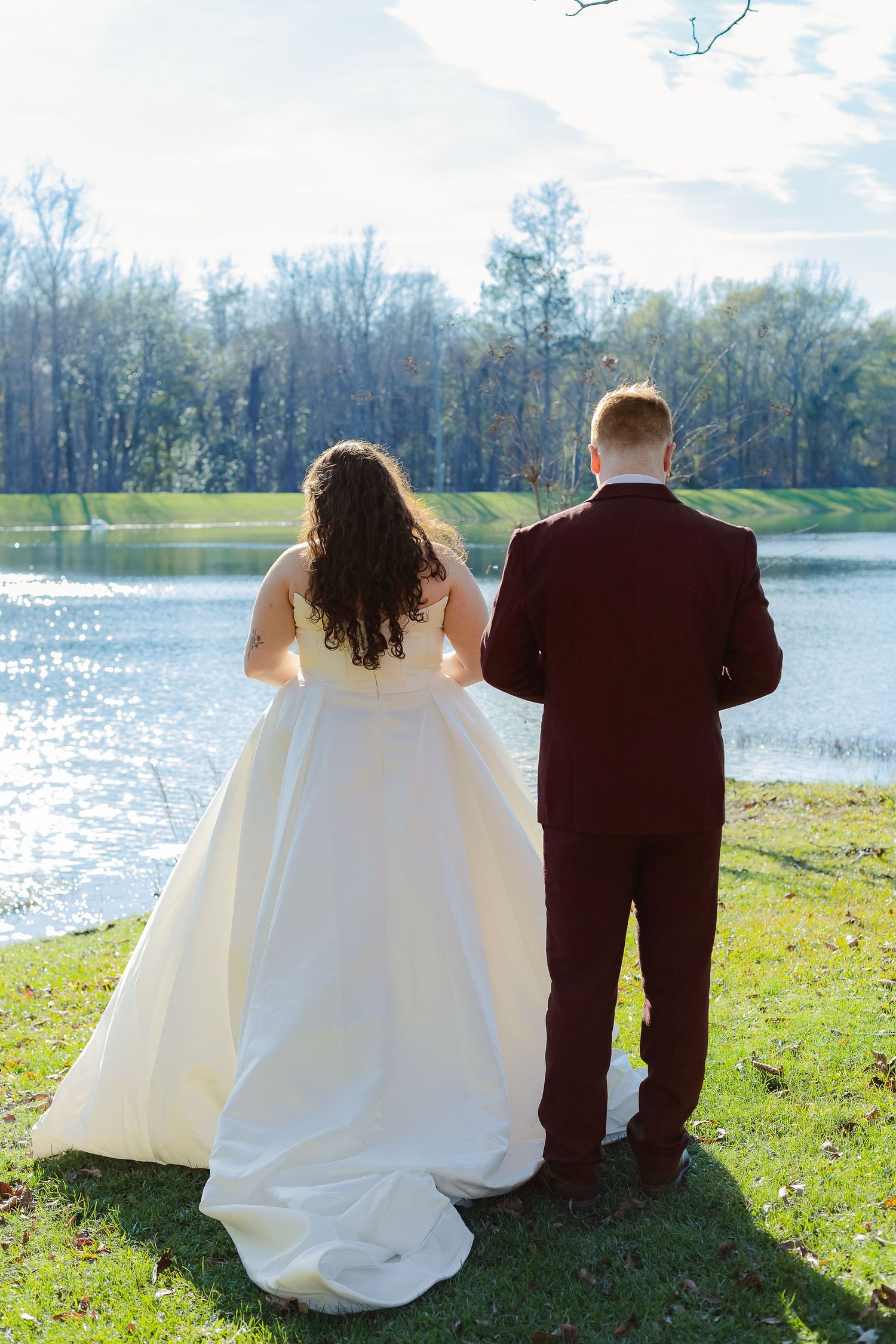 A couple sits together on grass by a serene lake, with the bride in white and groom in dark suit viewing the water.