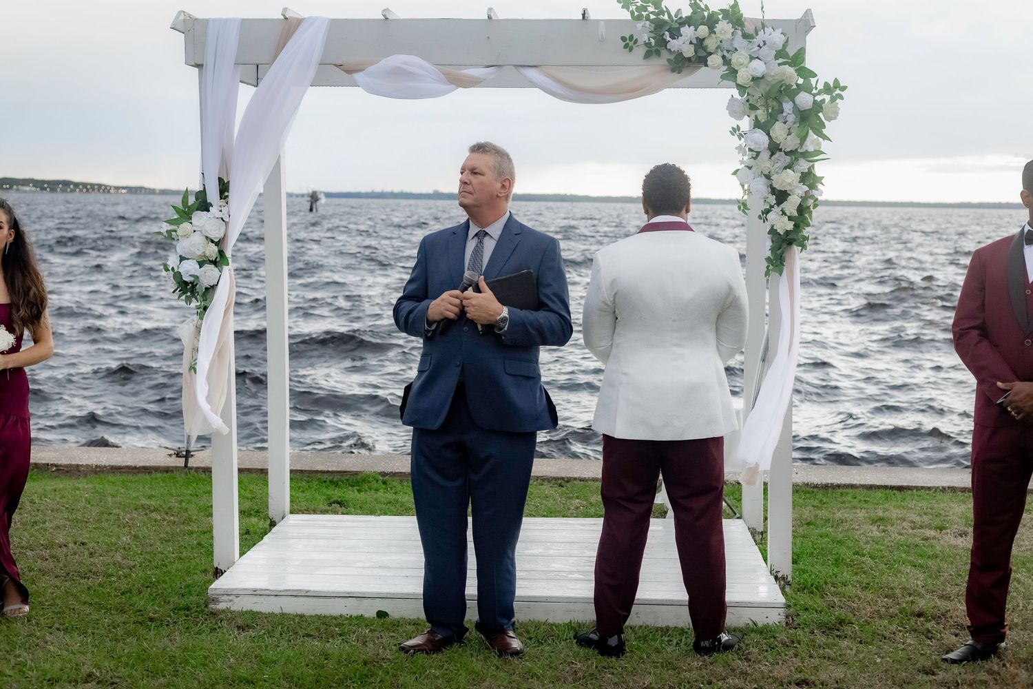 A white wedding arch decorated with floral arrangements stands on a grassy area overlooking a waterfront.
