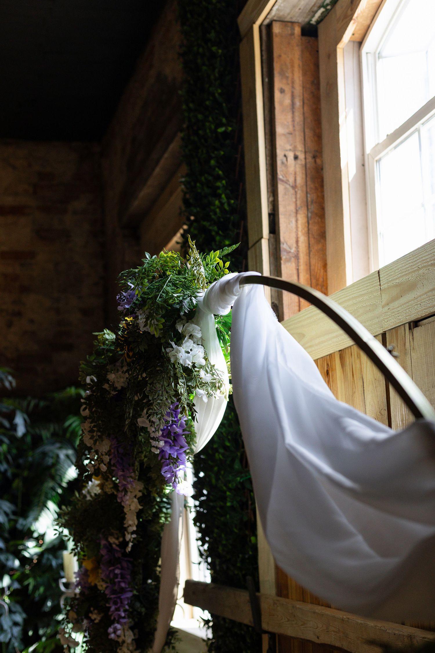 A white wedding dress hangs near a window with purple and green floral decorations in a rustic wooden setting.
