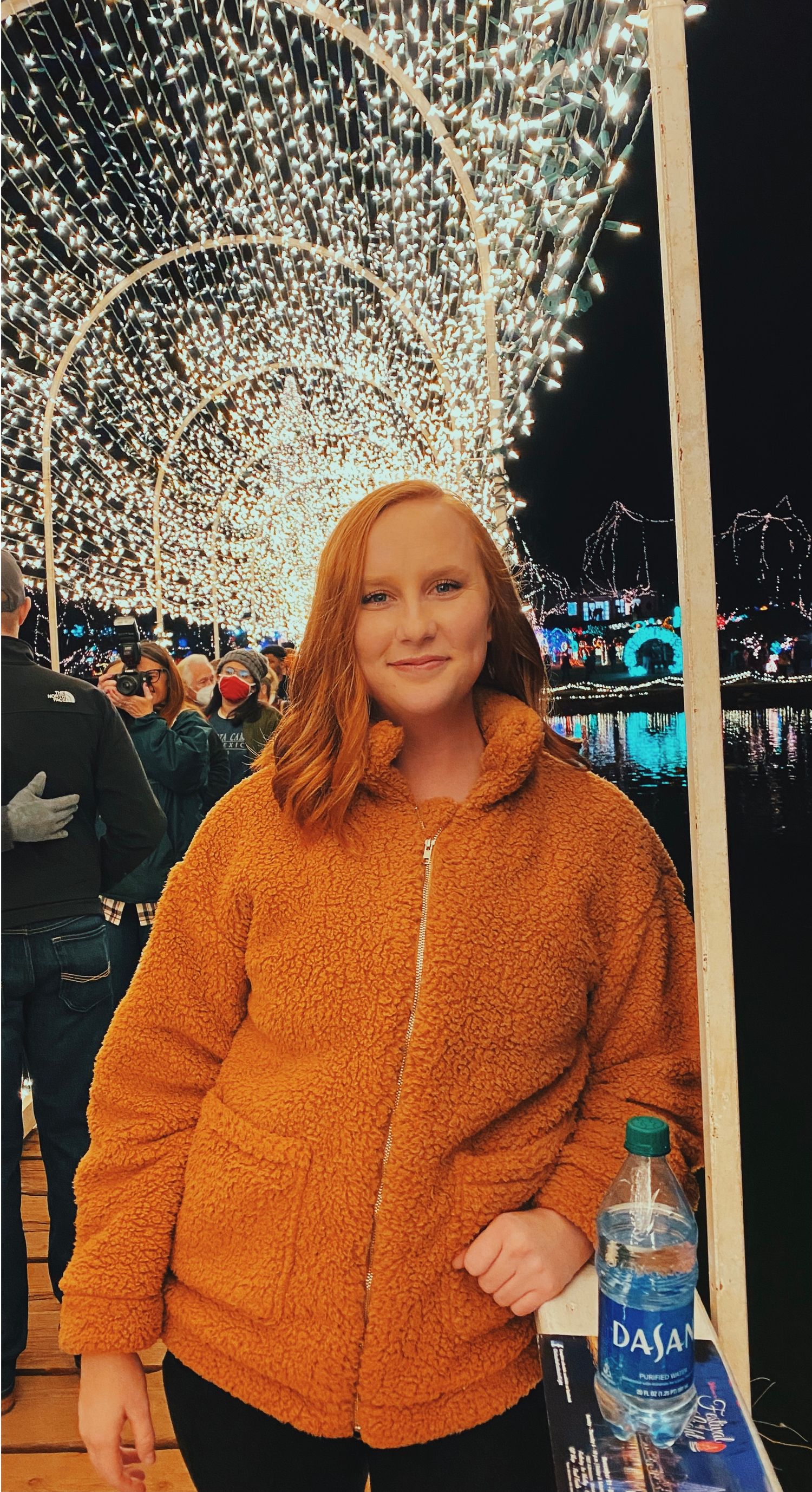 Person in orange fleece jacket standing under illuminated holiday light tunnel display at night.
