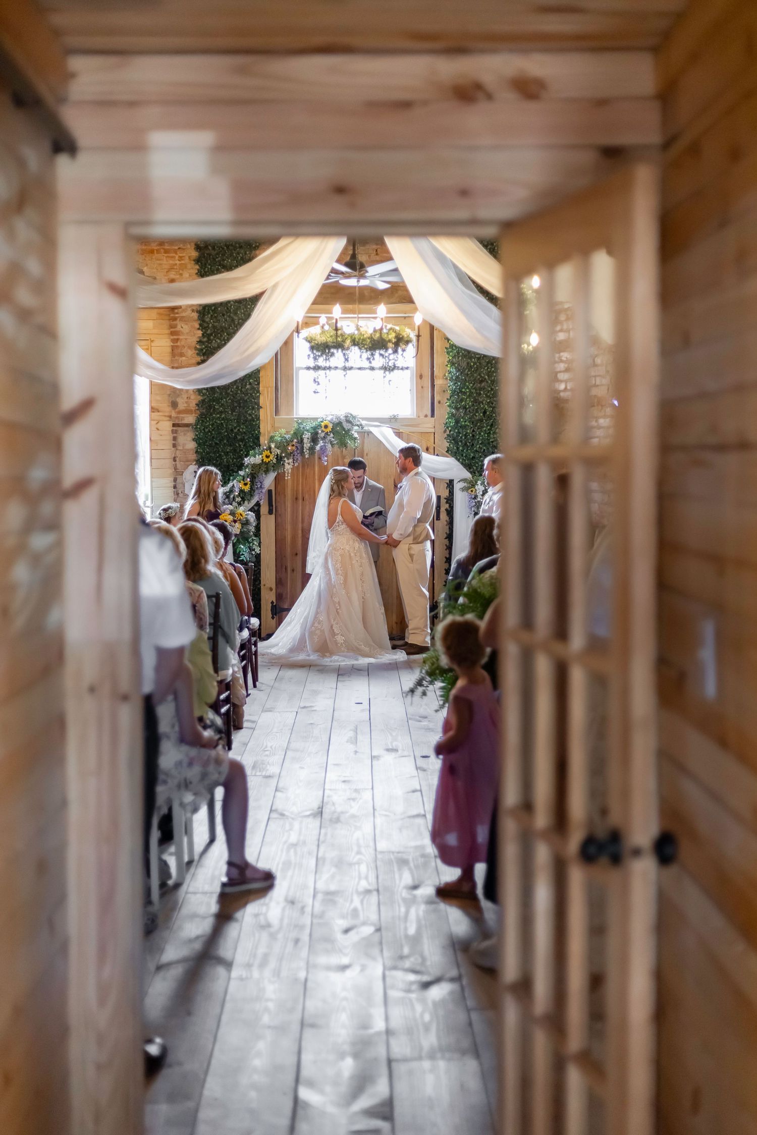 A wedding ceremony in a rustic wooden venue with white draping and guests lining the aisle.